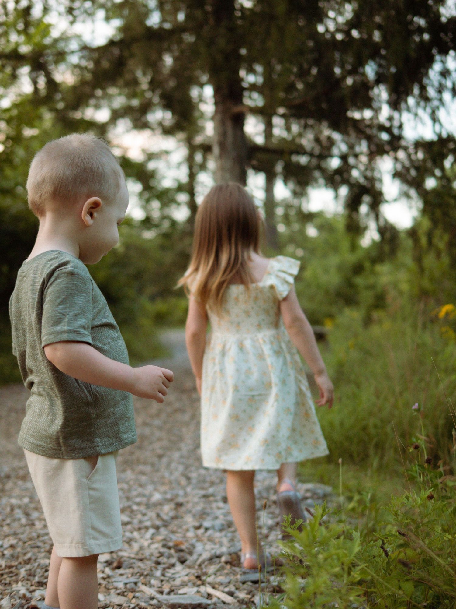 Two young children walk along a gravel garden path surrounded by lush green trees and wildflowers.