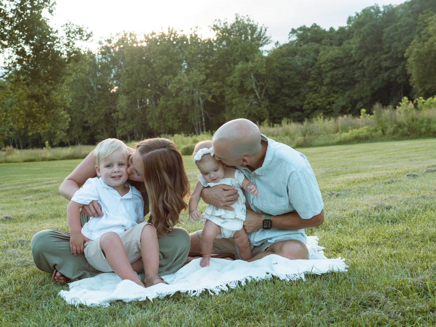 Happy interracial family of four sitting on white blanket in green meadow sharing kisses with children.