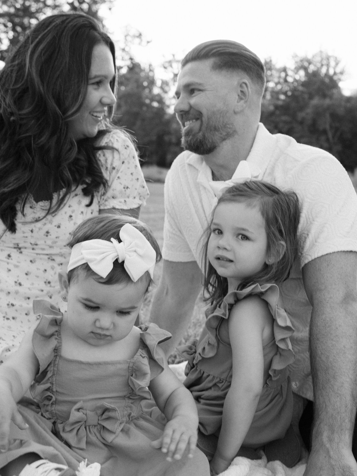 Black and white family portrait of parents smiling at each other with two young daughters outdoors.