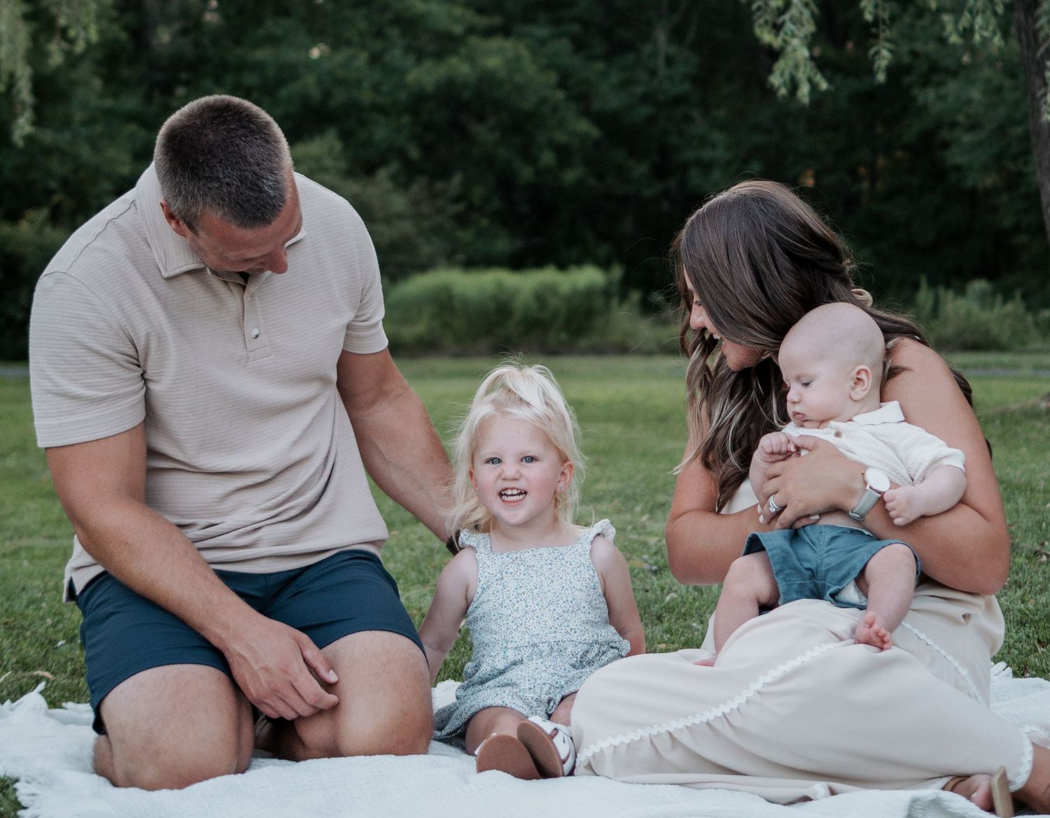 Family of four sits on blanket in park, parents smiling at toddler girl while mom holds newborn baby.