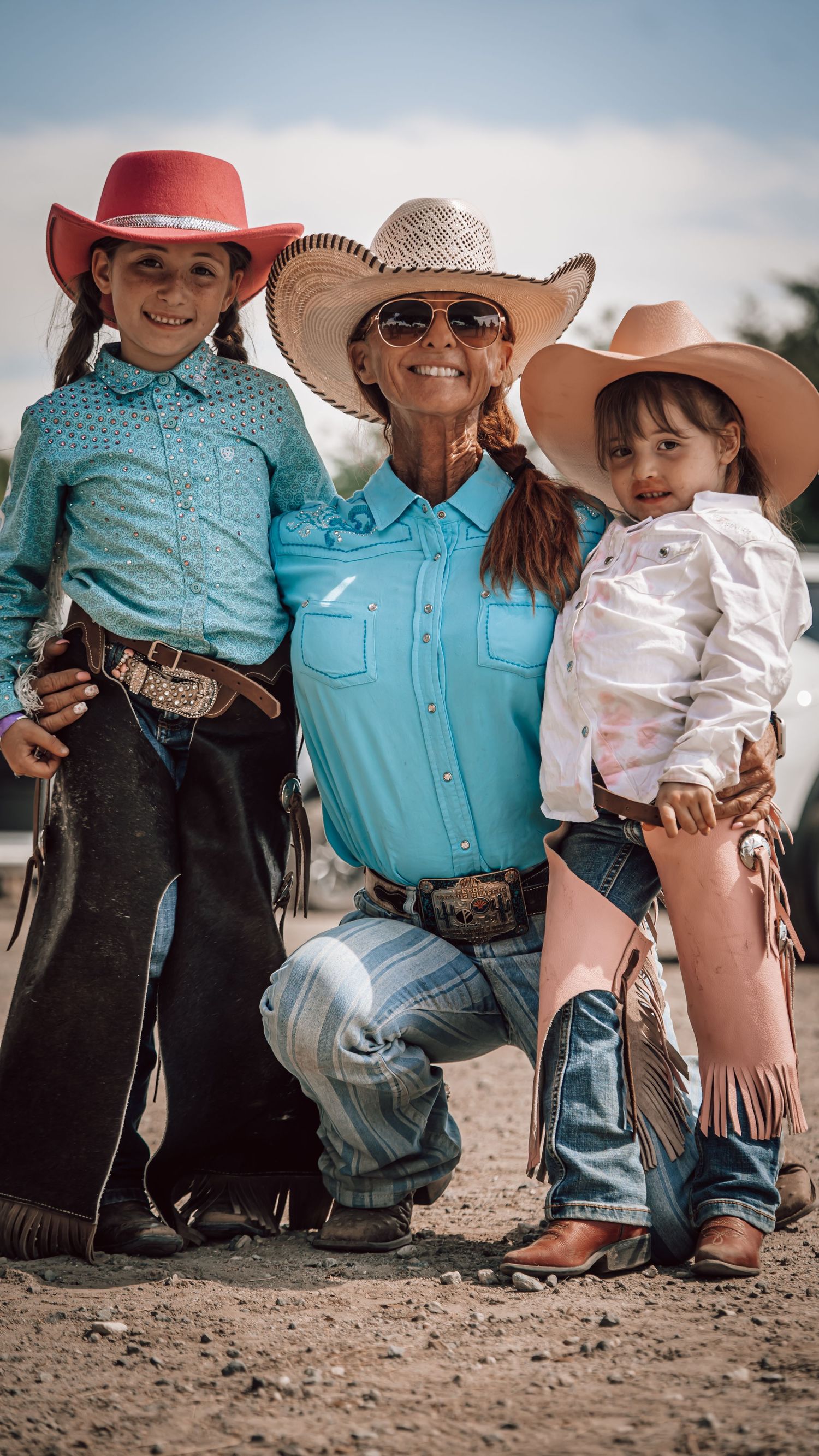 Three cowgirls in western attire, cowboy hats, and boots pose together outdoors at a rodeo event.