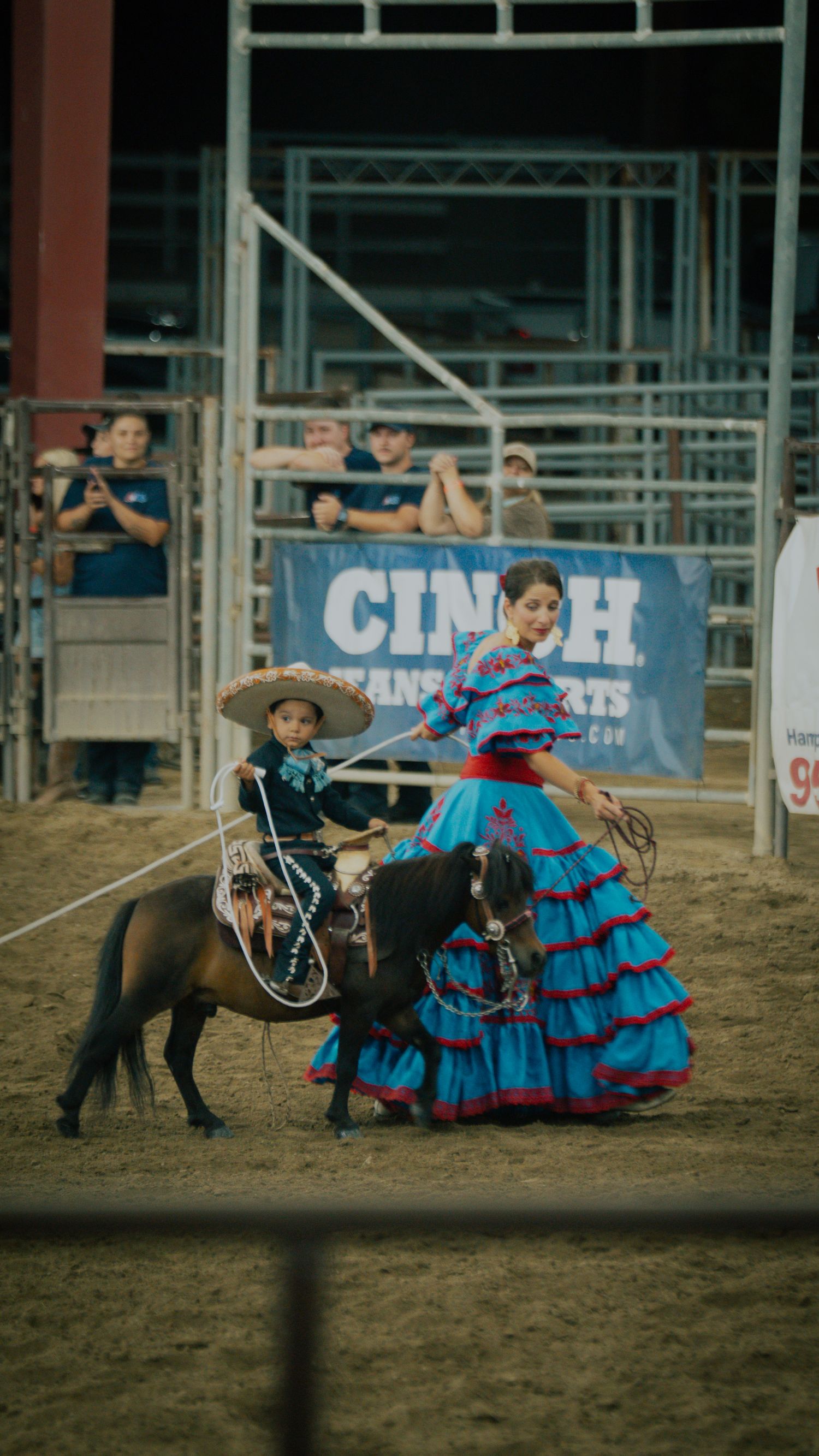 Child in sombrero rides pony beside woman in blue ruffled dress at a rodeo with CINCH banner behind them.
