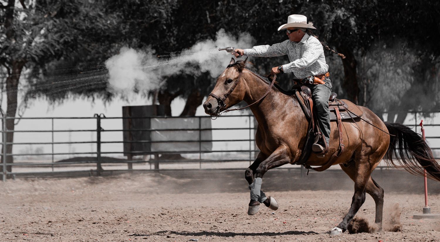 Cowboy mounted shooting competitor firing a revolver while riding a horse at a Western equestrian event