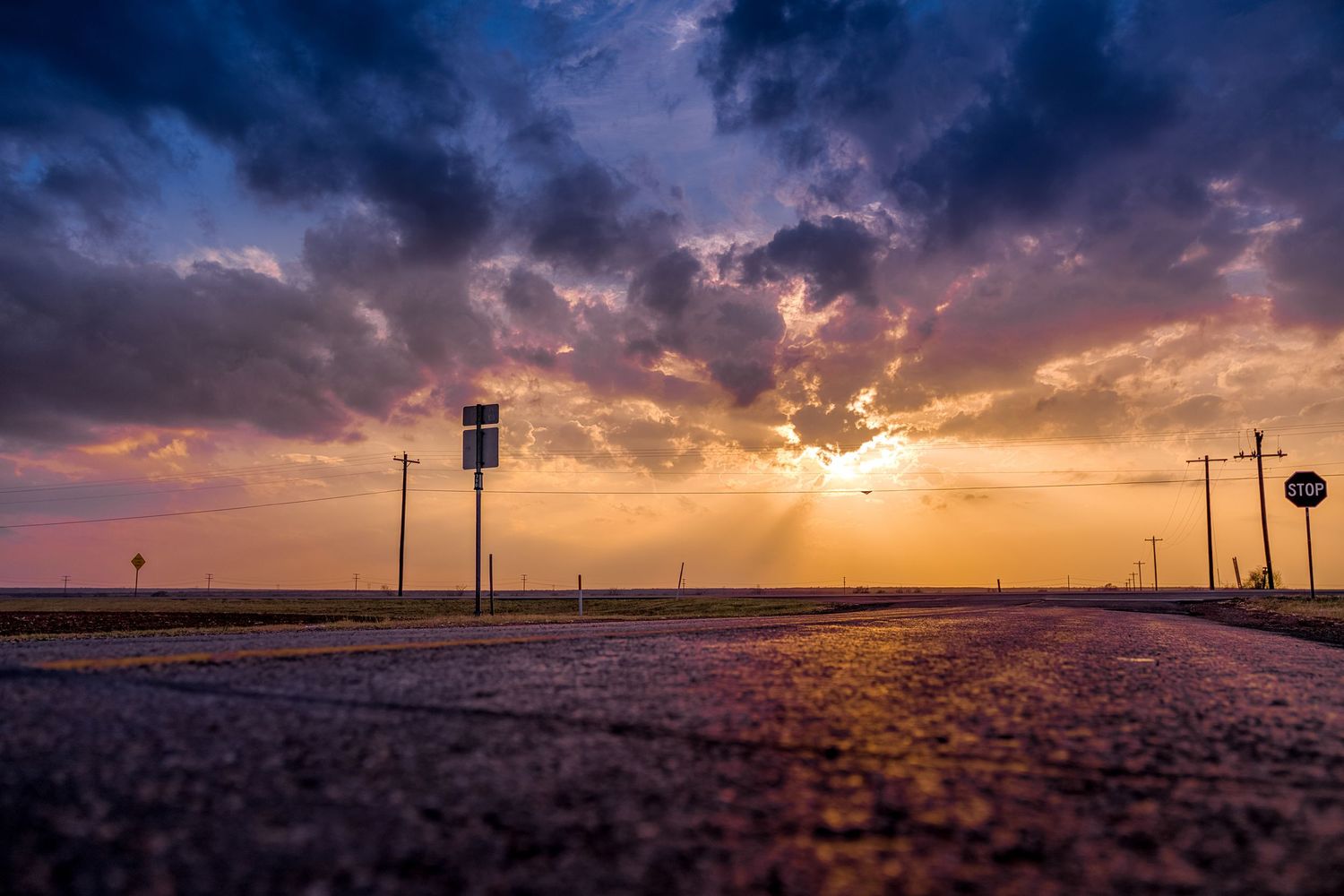 Golden sunlight breaks beneath storm clouds over a rural Texas Panhandle intersection with a water tower and utility poles.