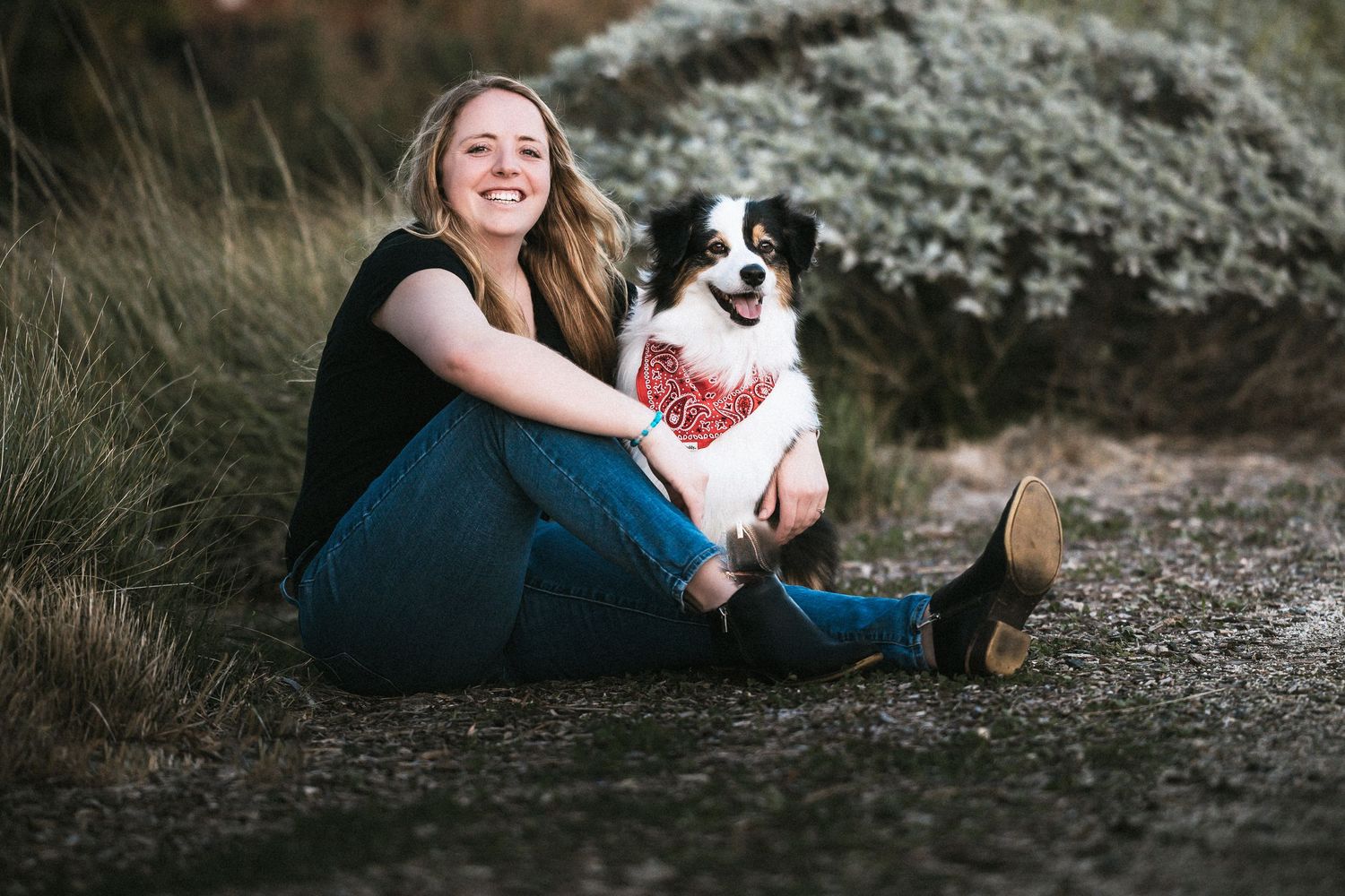Woman smiling and sitting outdoors with her border collie dog wearing a red bandana on a gravel path.