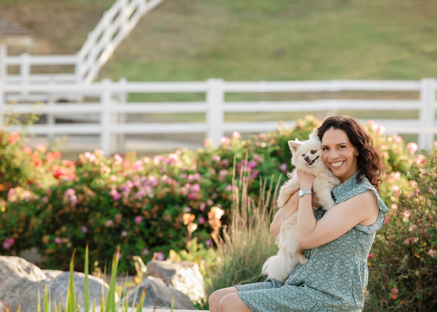 Woman in green dress smiling while holding a white puppy outdoors near a white fence and colorful flower garden.