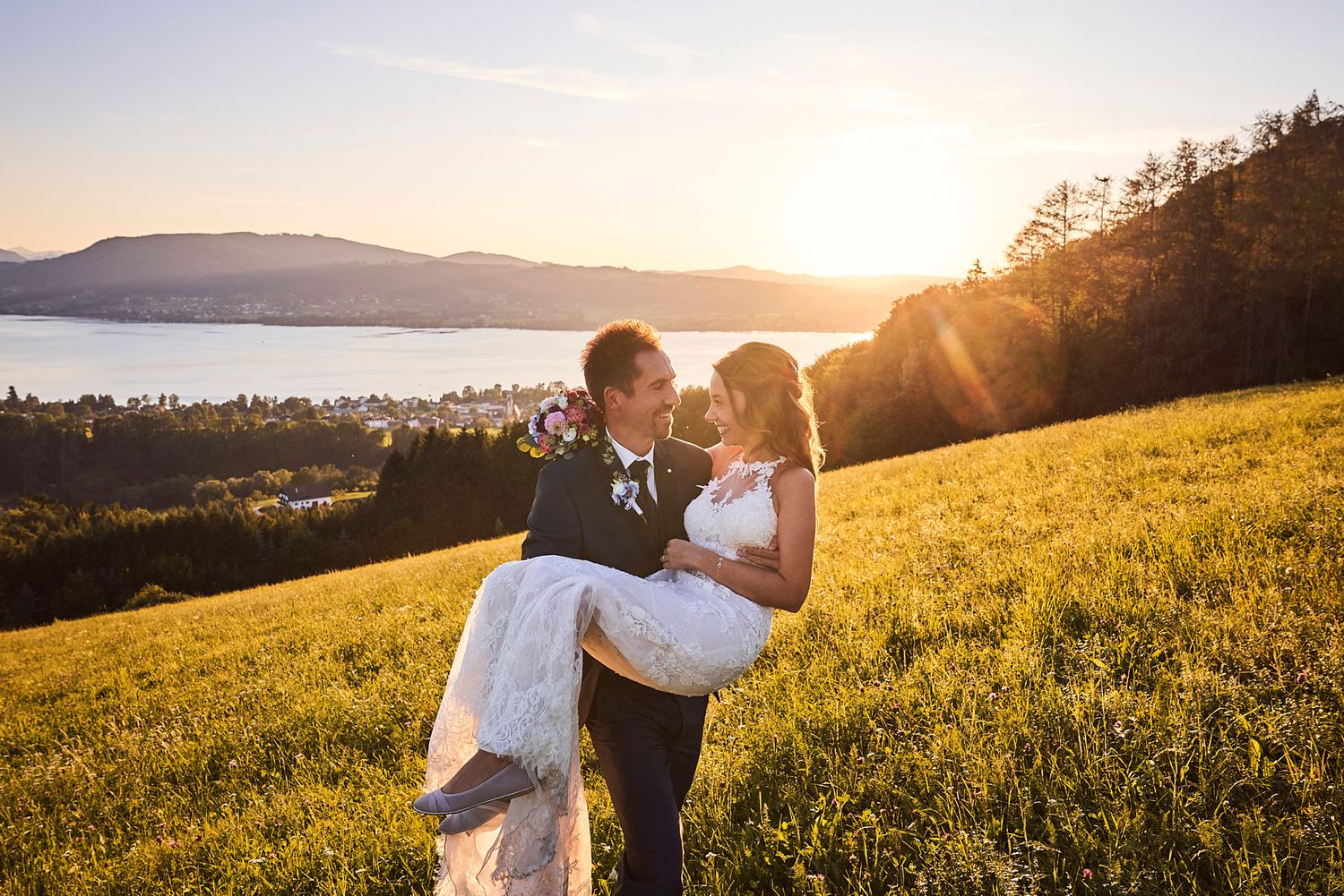 Marié portant la mariée dans un champ au coucher du soleil, au bord d'un lac en Autriche