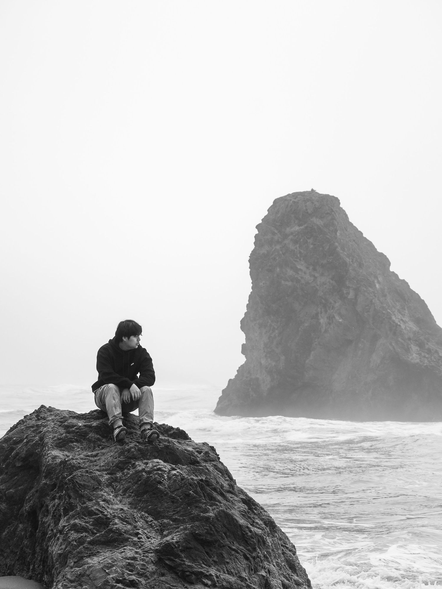 Black-and-white photograph of a person sitting on coastal rocks with a sea stack in the distance.