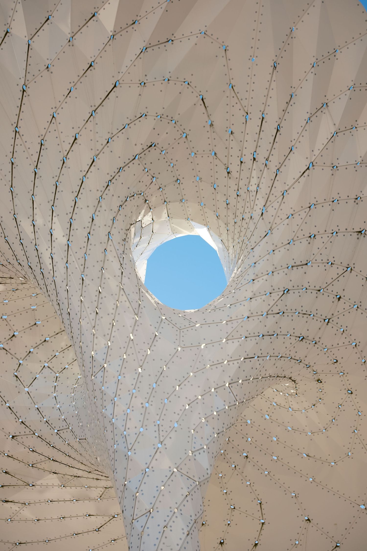 Looking upward through a geometric white architectural structure toward a blue sky.