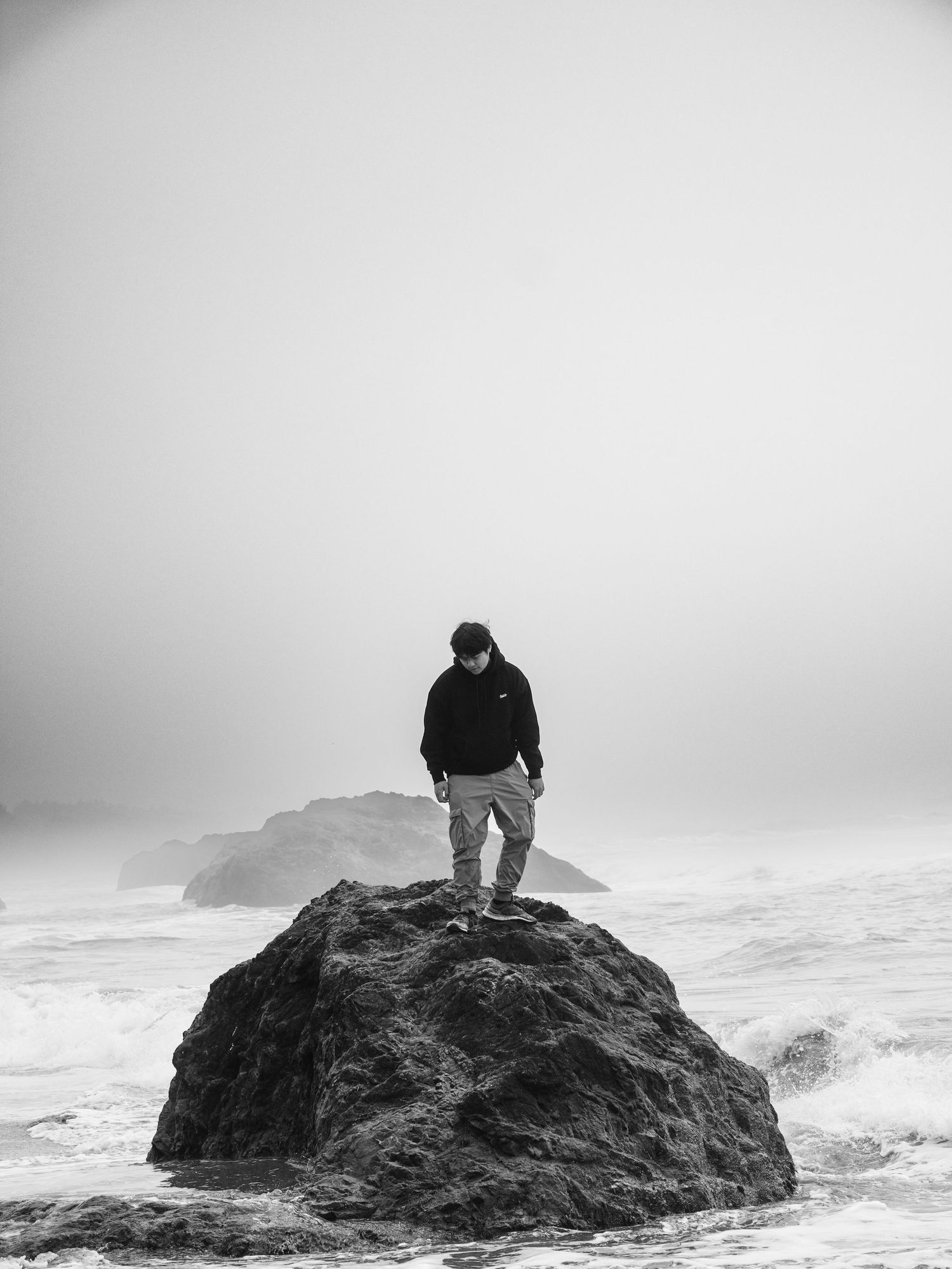 Black-and-white photograph of a person standing atop a rock in the ocean.