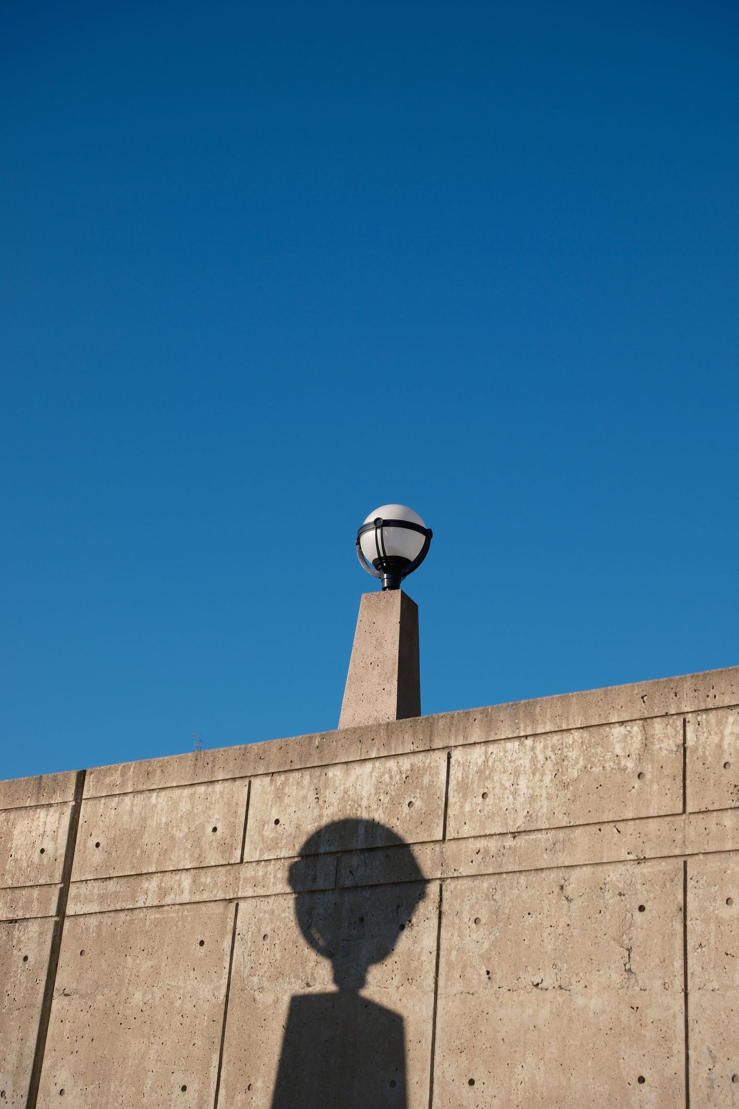 Minimal architectural photograph of a light fixture and its shadow against a concrete wall and blue sky.