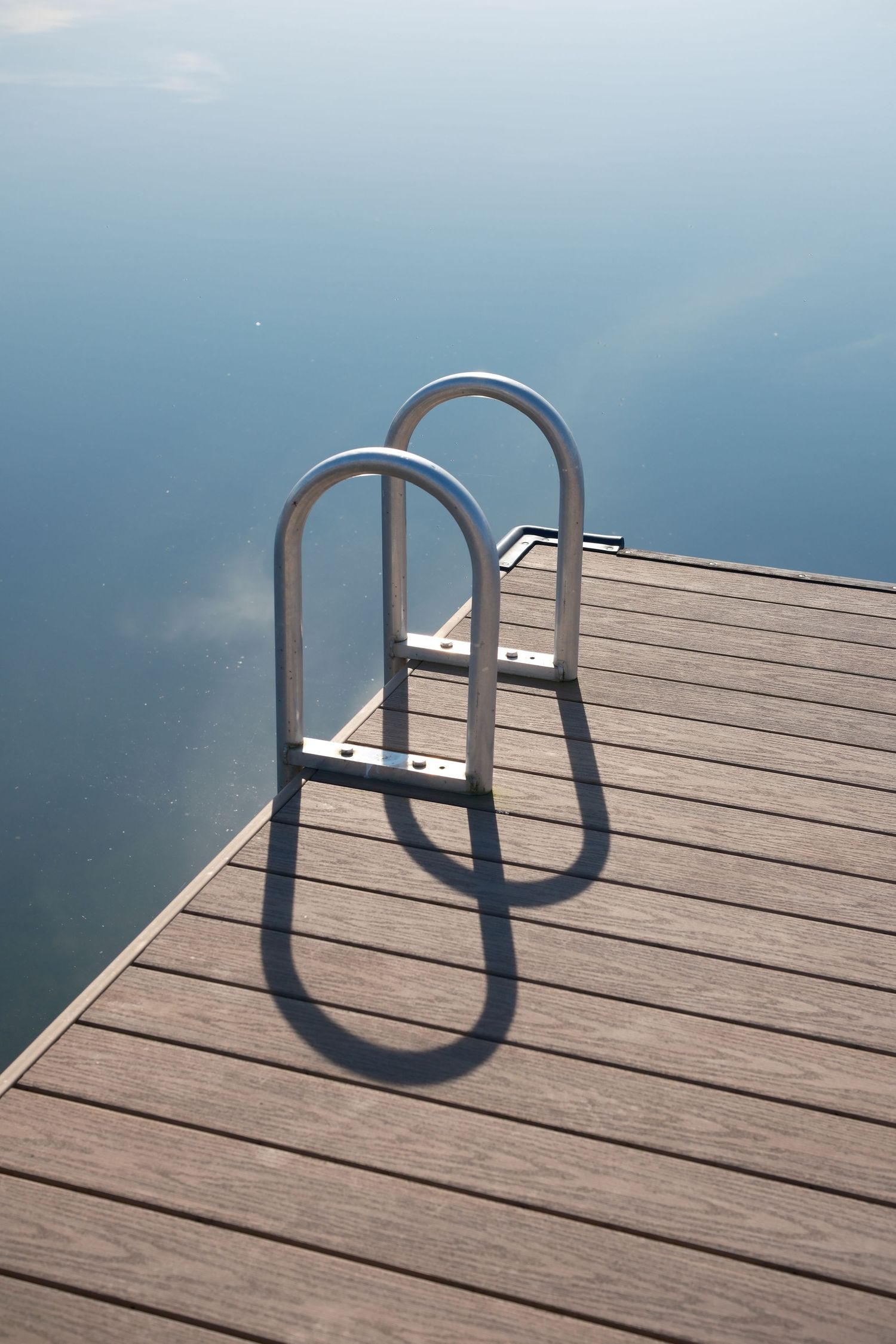 Metal pool ladder casting a shadow across a wooden dock beside calm water.