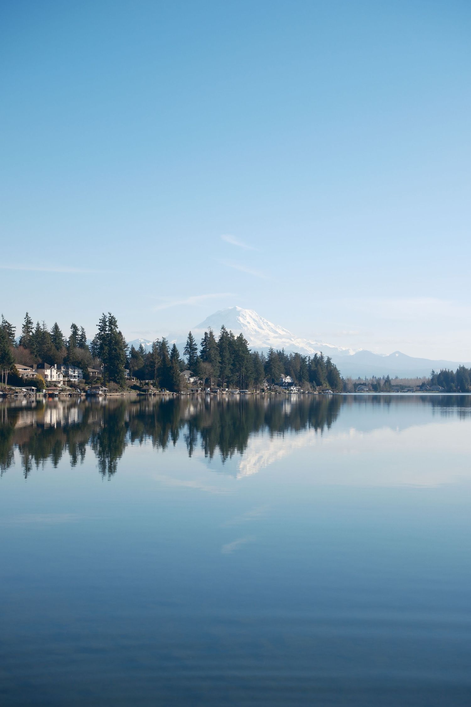 Calm waterfront landscape with houses and Mount Rainier reflected in the water beneath a clear blue sky.