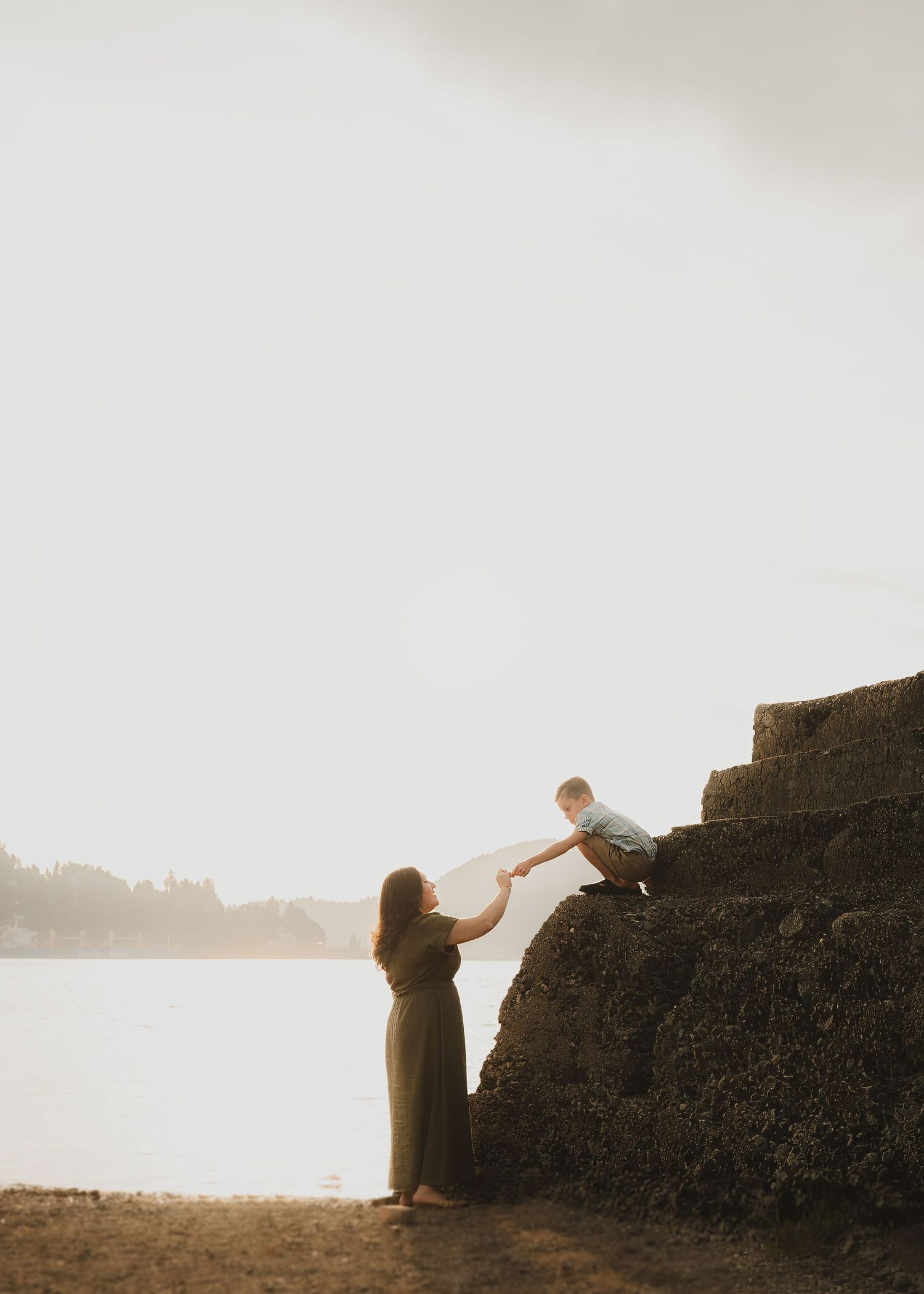 Woman in a long olive dress holds the hand of a young boy climbing a rocky ledge beside a calm lake at soft golden sunset.