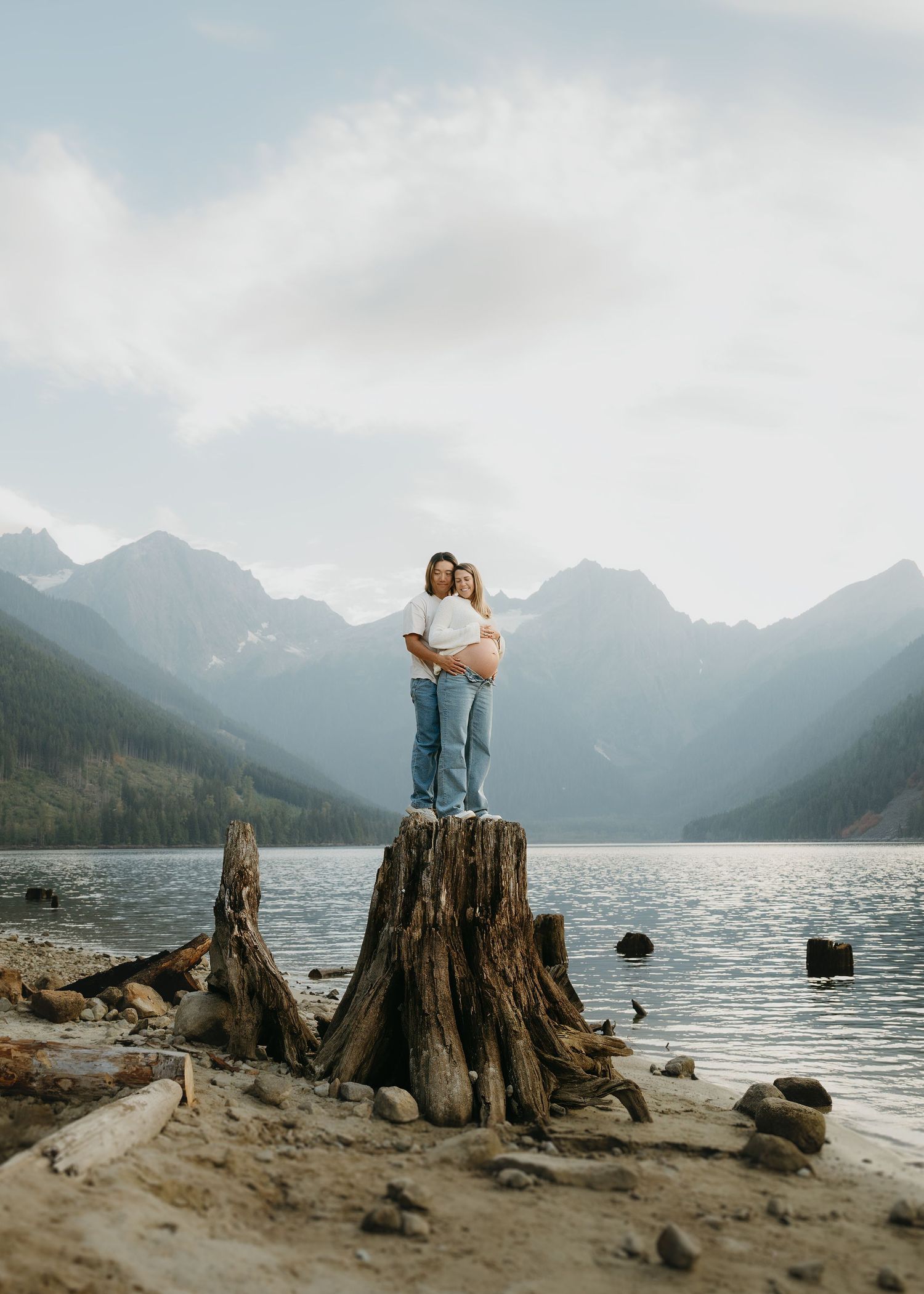 A couple stands on a tree stump overlooking a serene mountain lake with misty peaks in the background.