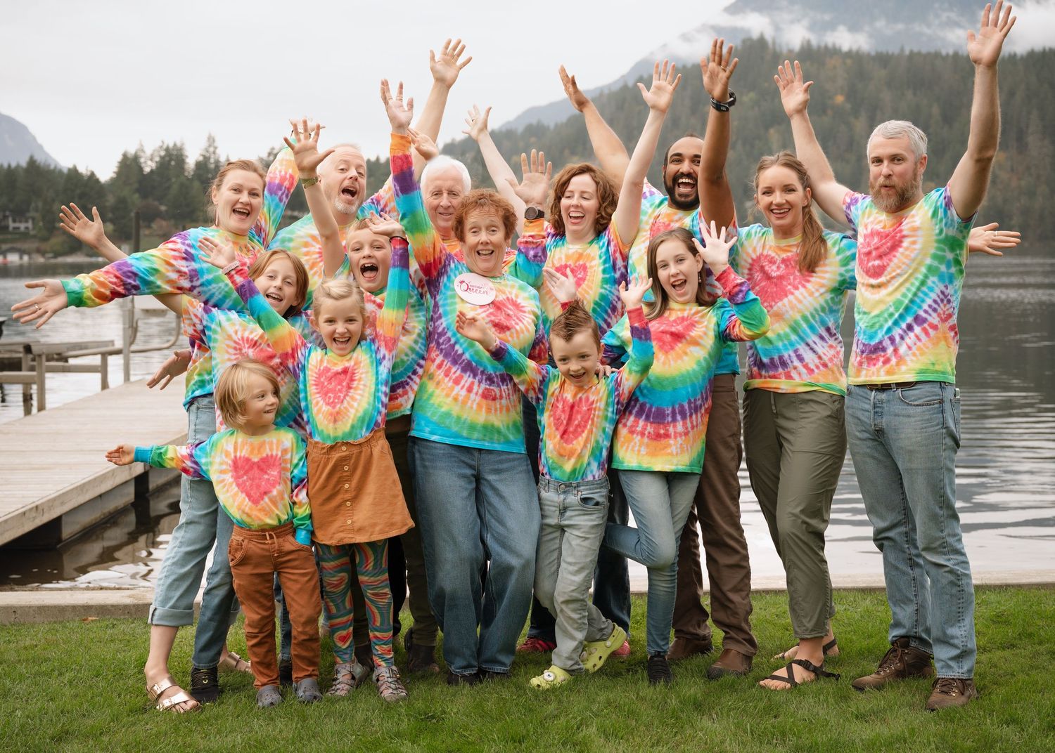 A cheerful group wearing colorful tie-dye shirts poses with raised arms by a lakeside dock with mountains in the background.