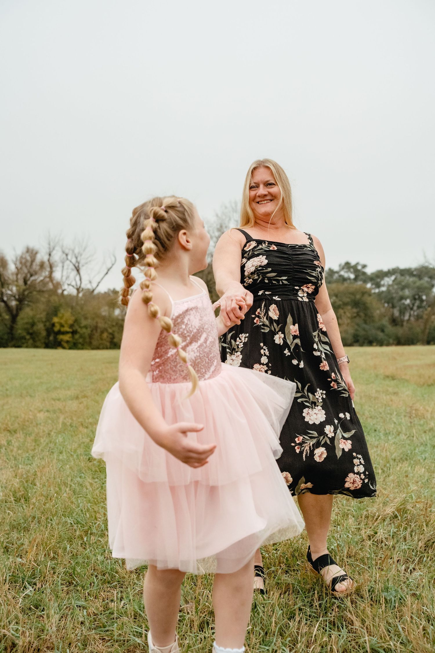 A smiling blonde woman in a floral dress holds hands with a young girl in a pink tutu dress in a grassy field.