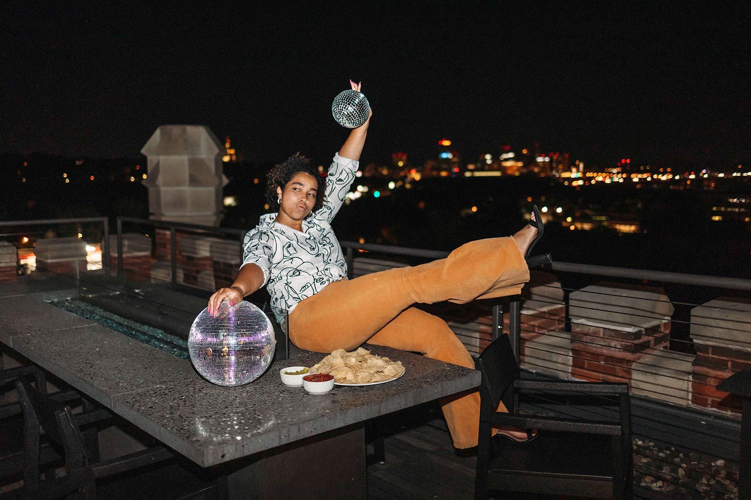 Evening rooftop setting with disco ball and city lights in background.