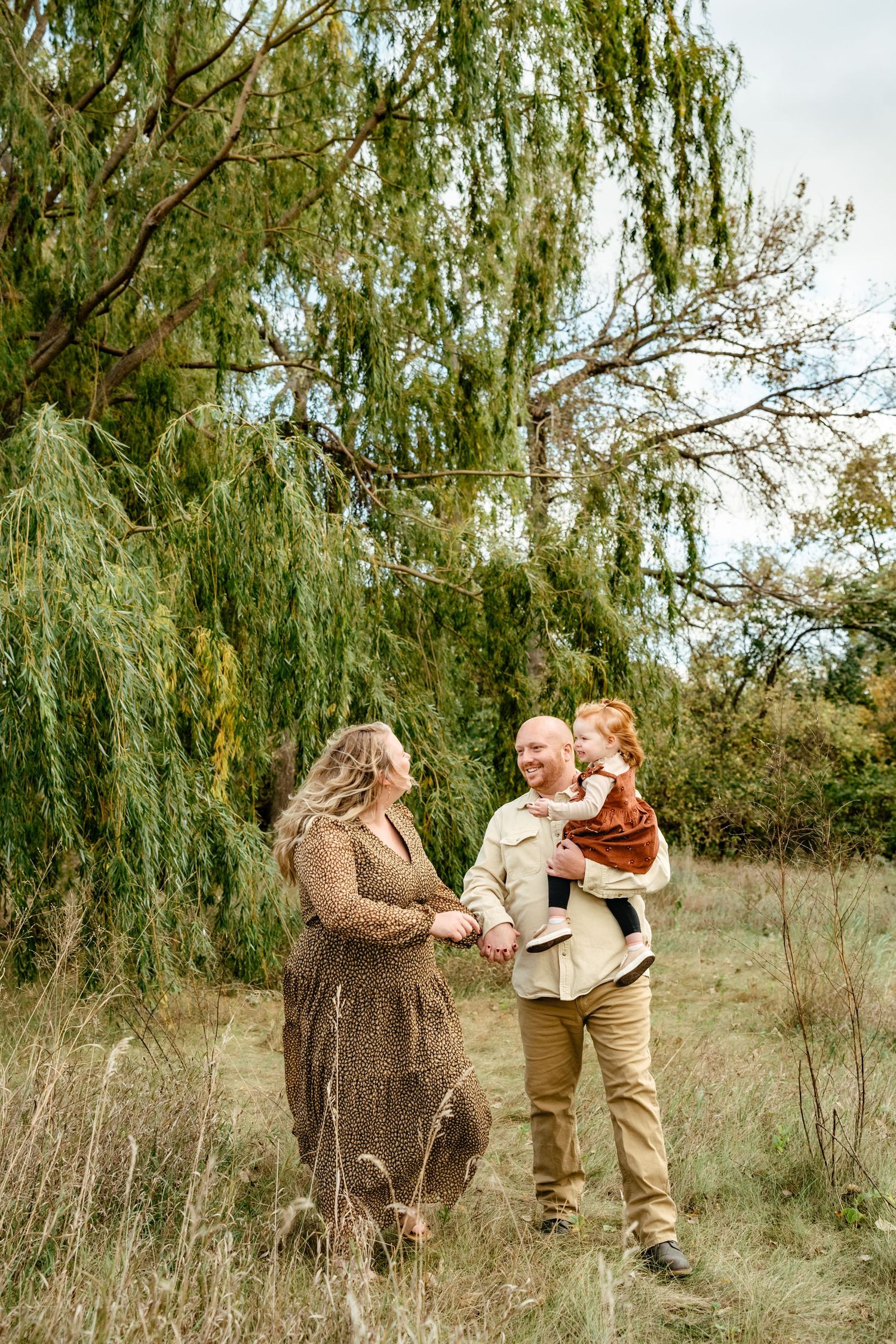 A family of three poses outdoors near a weeping willow, parents holding hands with their red-haired toddler in autumn.