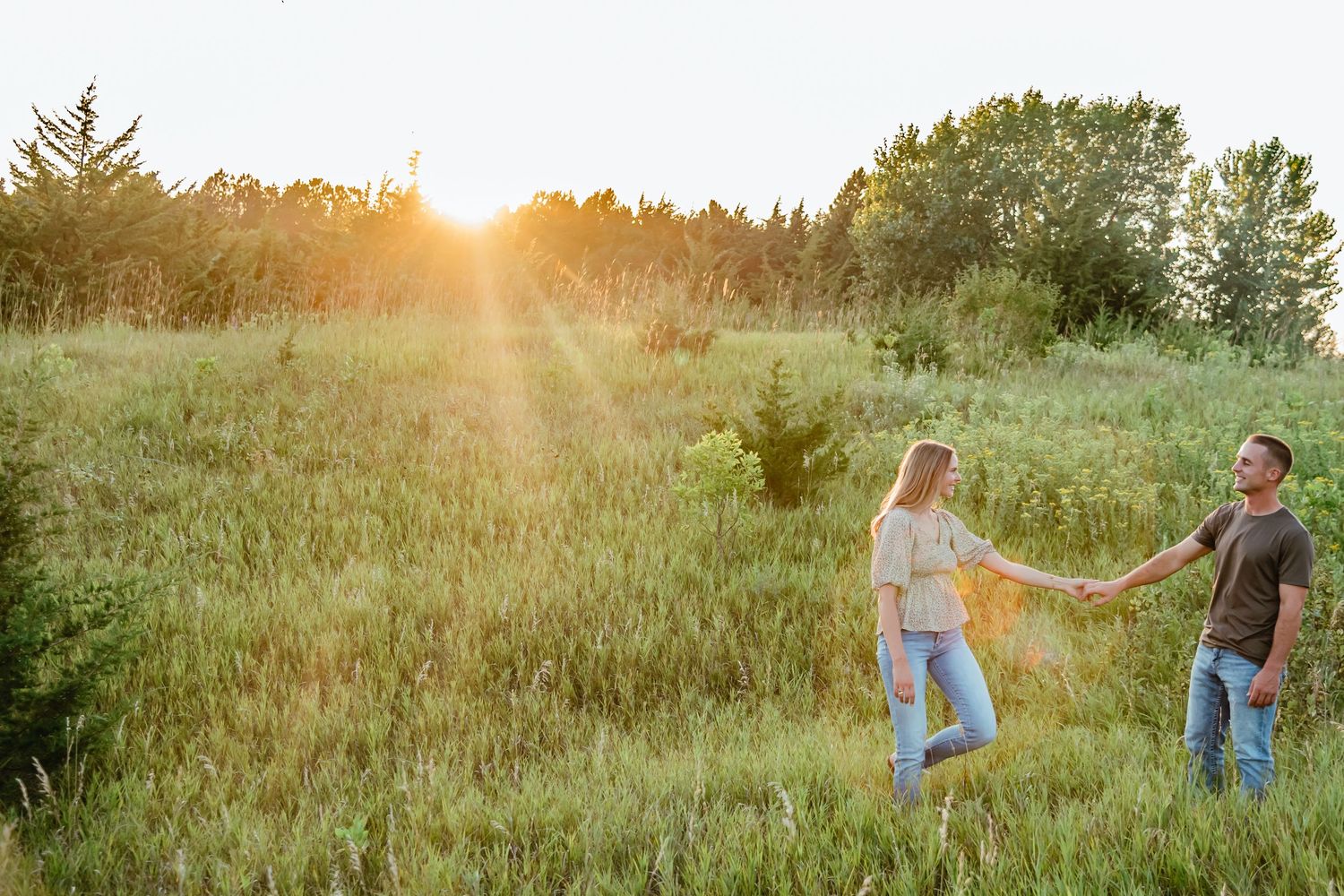 Couple holding hands walking through a sunlit green meadow at golden hour with trees in the background.