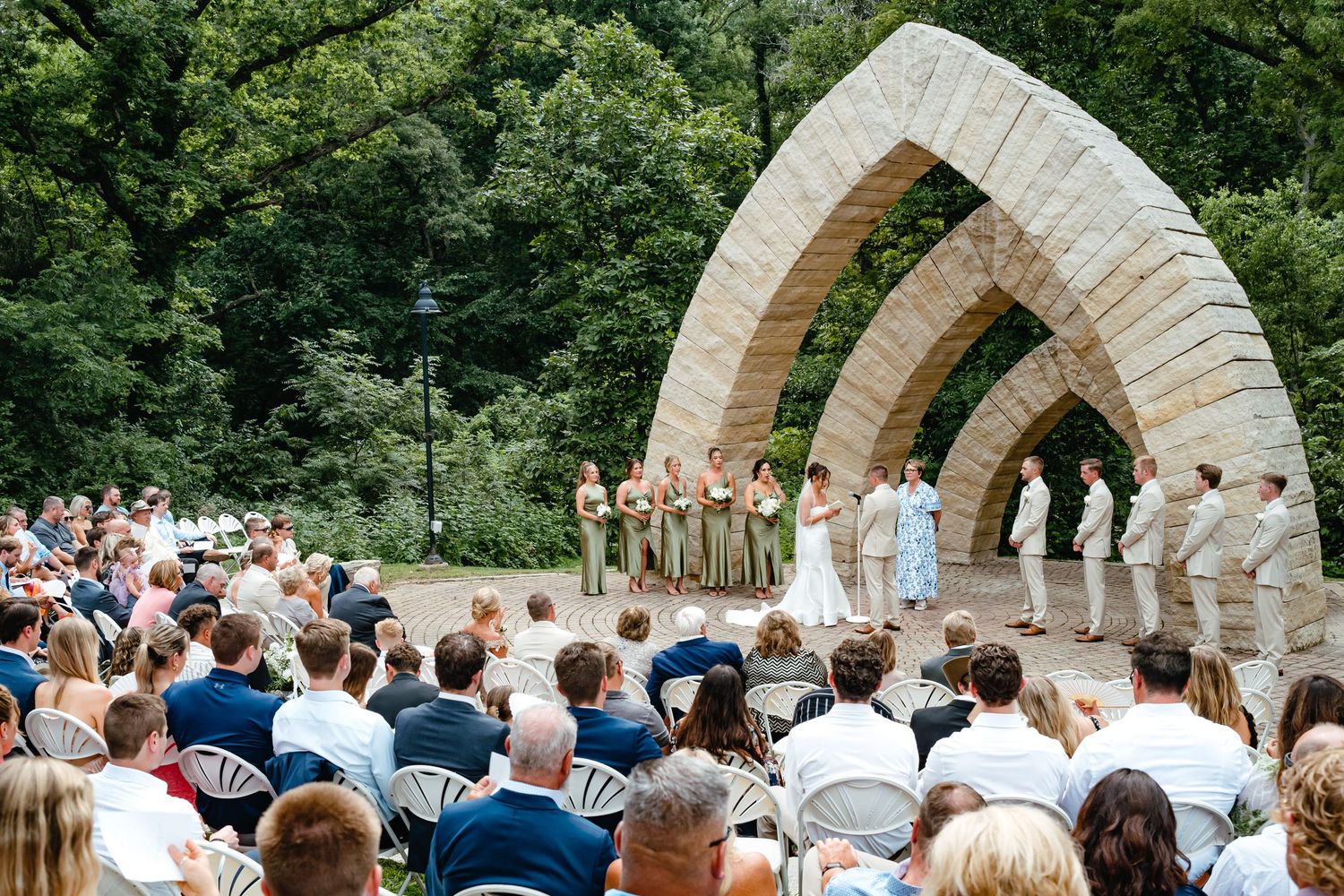 Wedding ceremony under stone archways with guests seated and greenery in background.