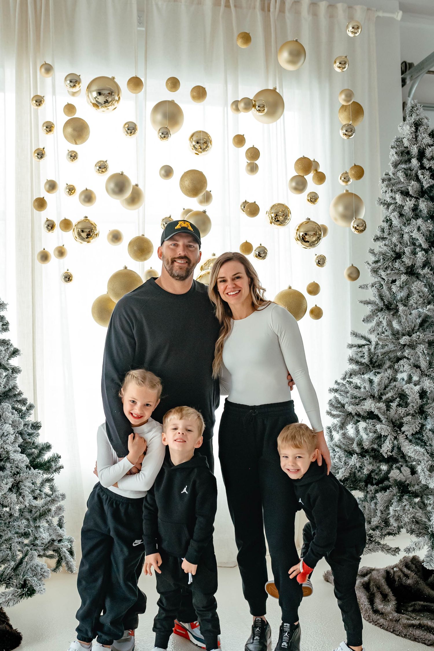 Family poses together in coordinated black and white outfits against white Christmas backdrop with gold ornaments.