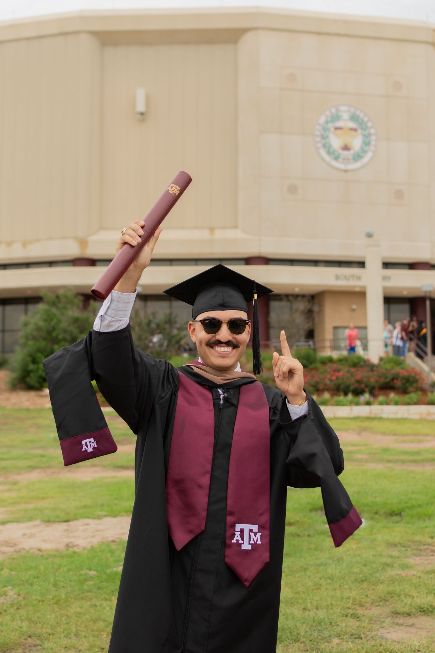 An Aggie Masters grad holds his diploma with one hand & his pointer finger on the other hand to give thanks to God while smiling at family.