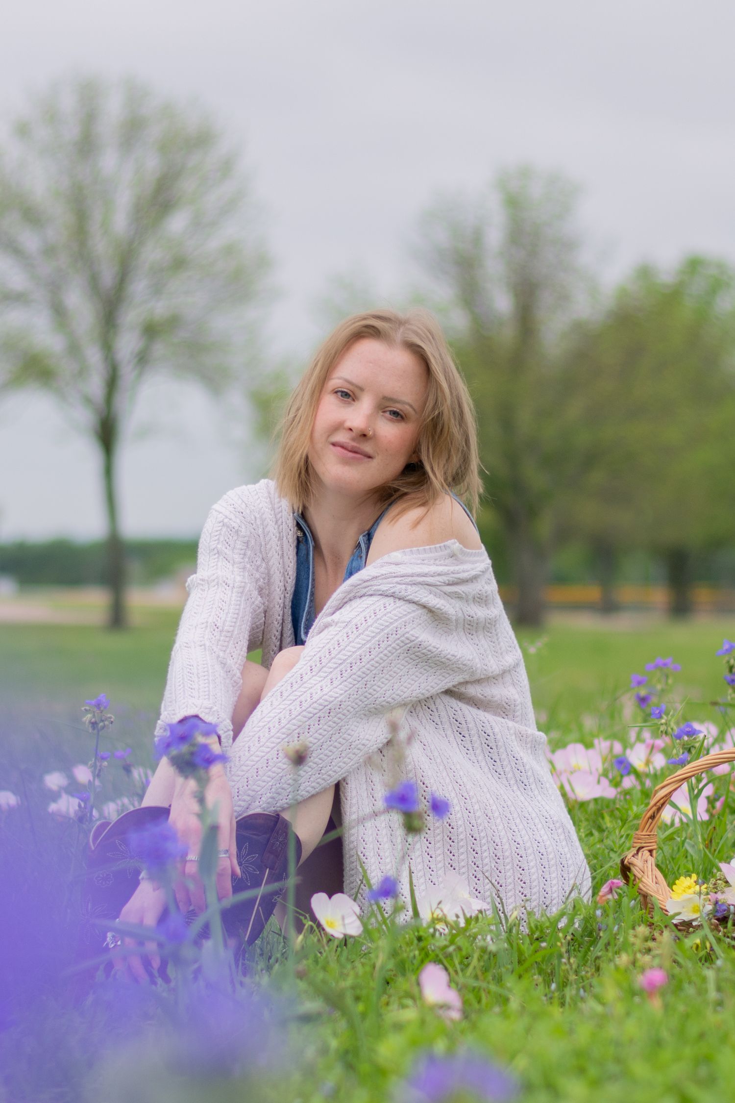 A young woman sits in a flower field on a cloudy spring day in Fort Worth, Texas for a portrait. She softly smiles with her head tilted.