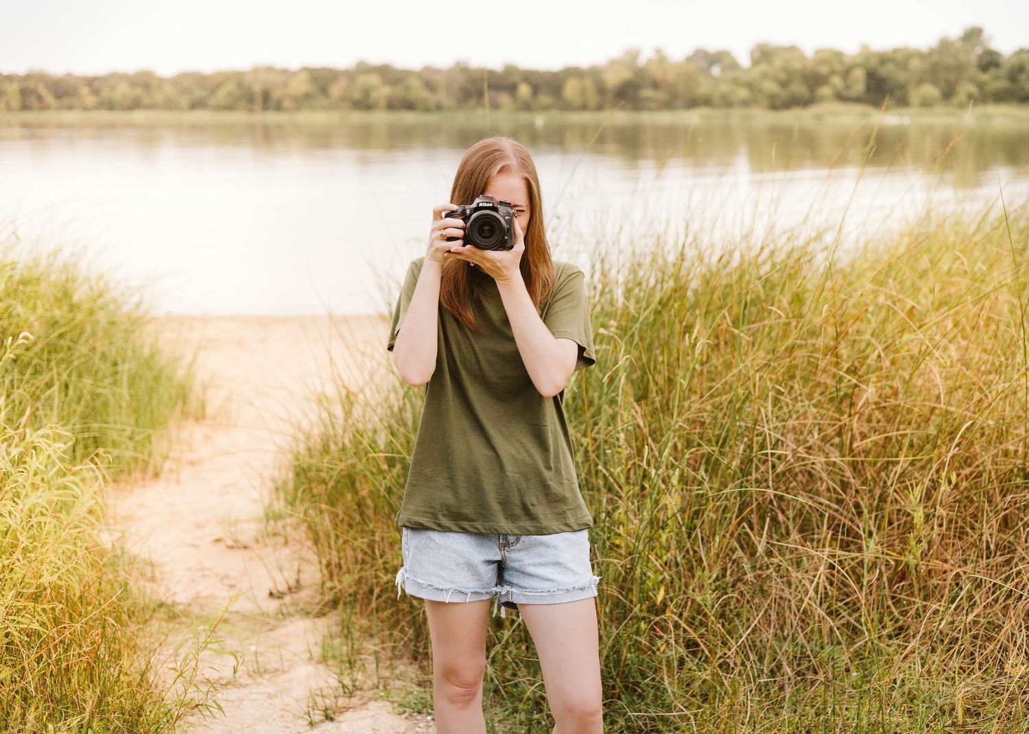A photo of Brooke, owner of Brooke Gonzalez Photography, standing near tall grass & a lake on a sunny day in Fort Worth, holding her camera.