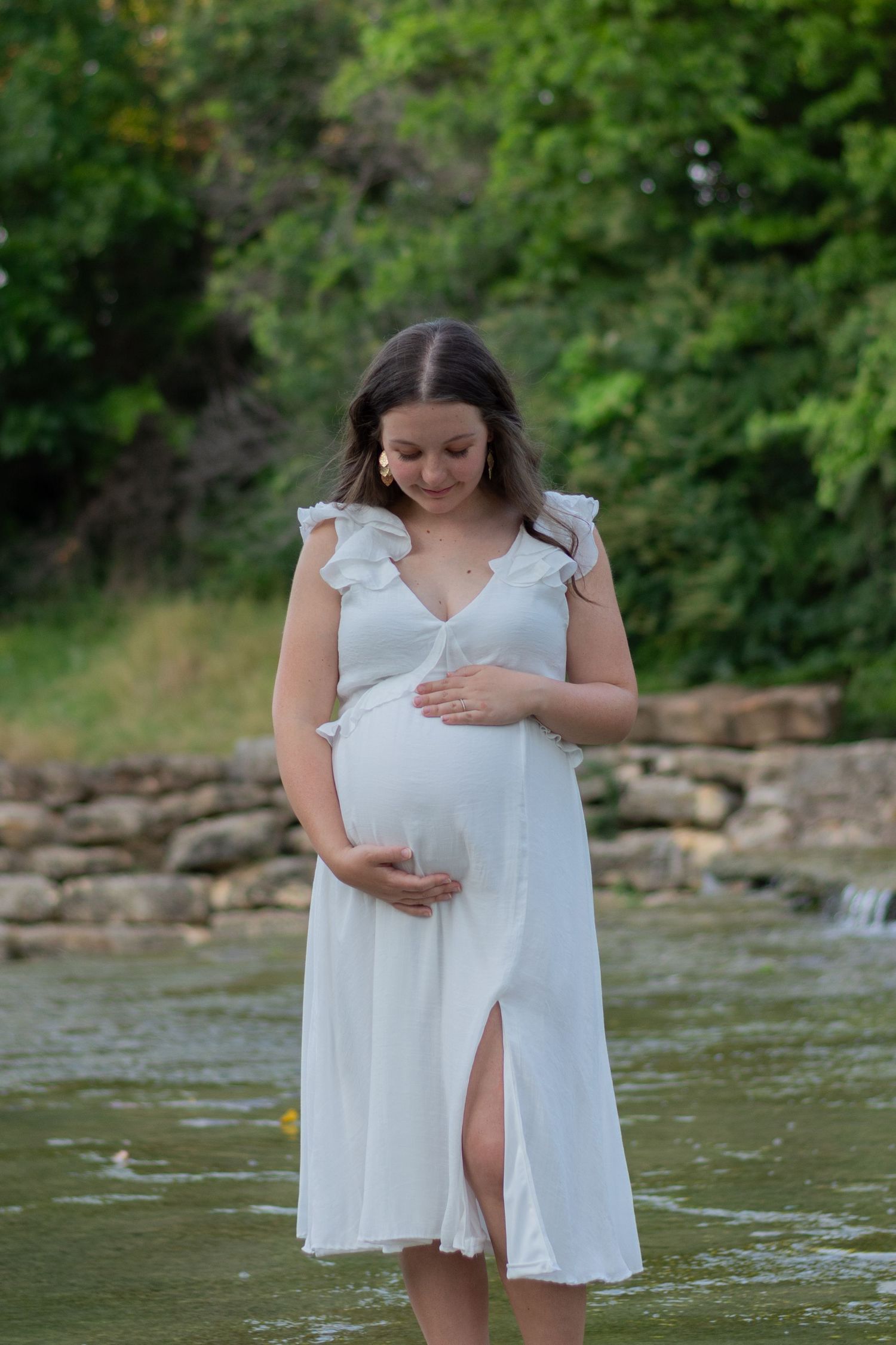A pregnant woman in a white flowy dress holds her belly while looking down. She's standing in a river in Fort Worth, TX in the summer.