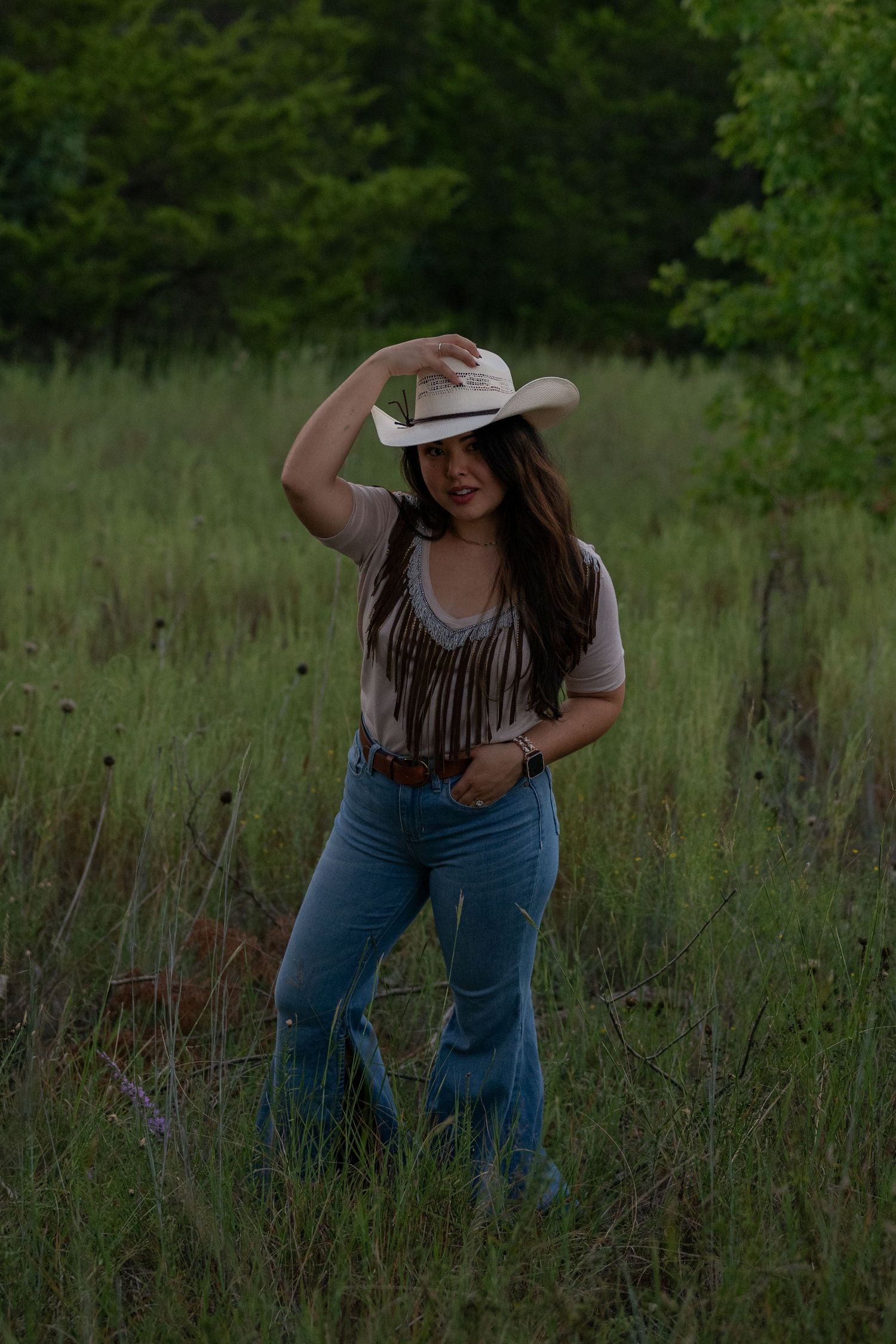 A woman poses in a grassy field while dressed in a western outfit with fringe & cowgirl hat, holding her hat with one hand, other in pocket.