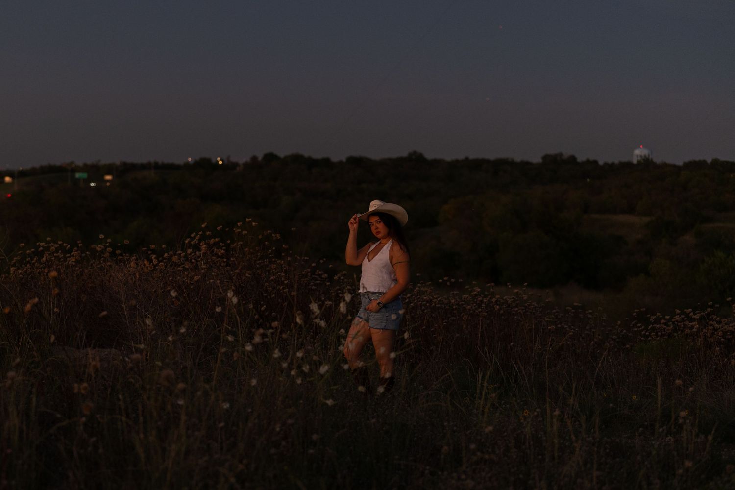 A woman poses in a field during blue hour at a creative portrait shoot. She's wearing boots, denim shorts, a white halter, & touches her hat