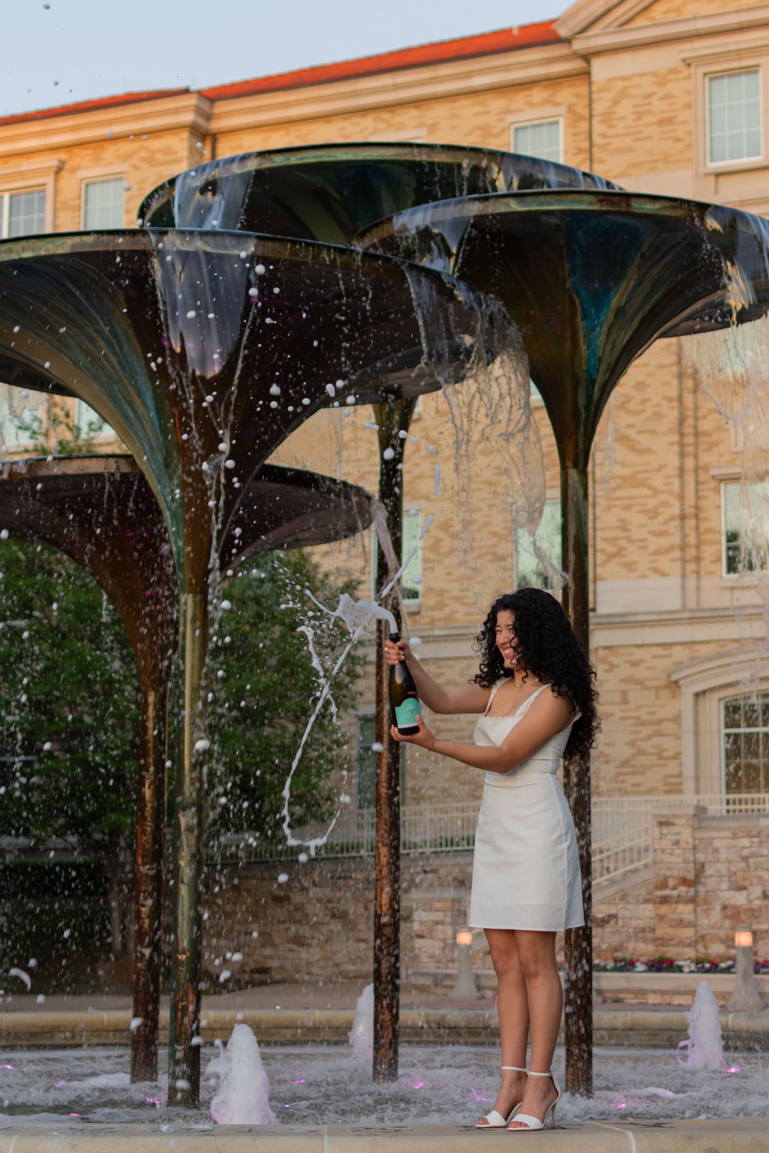A Texas Christian University grad pops a bottle of champagne joyfully while standing on Frog Fountain in her white dress and heels.