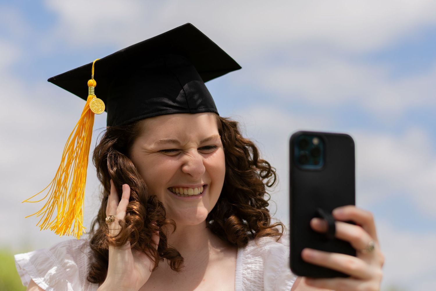 Candid photo of a smiling grad in a white dress and grad cap with yellow tassel taking a selfie during a grad photoshoot to send to her mom.