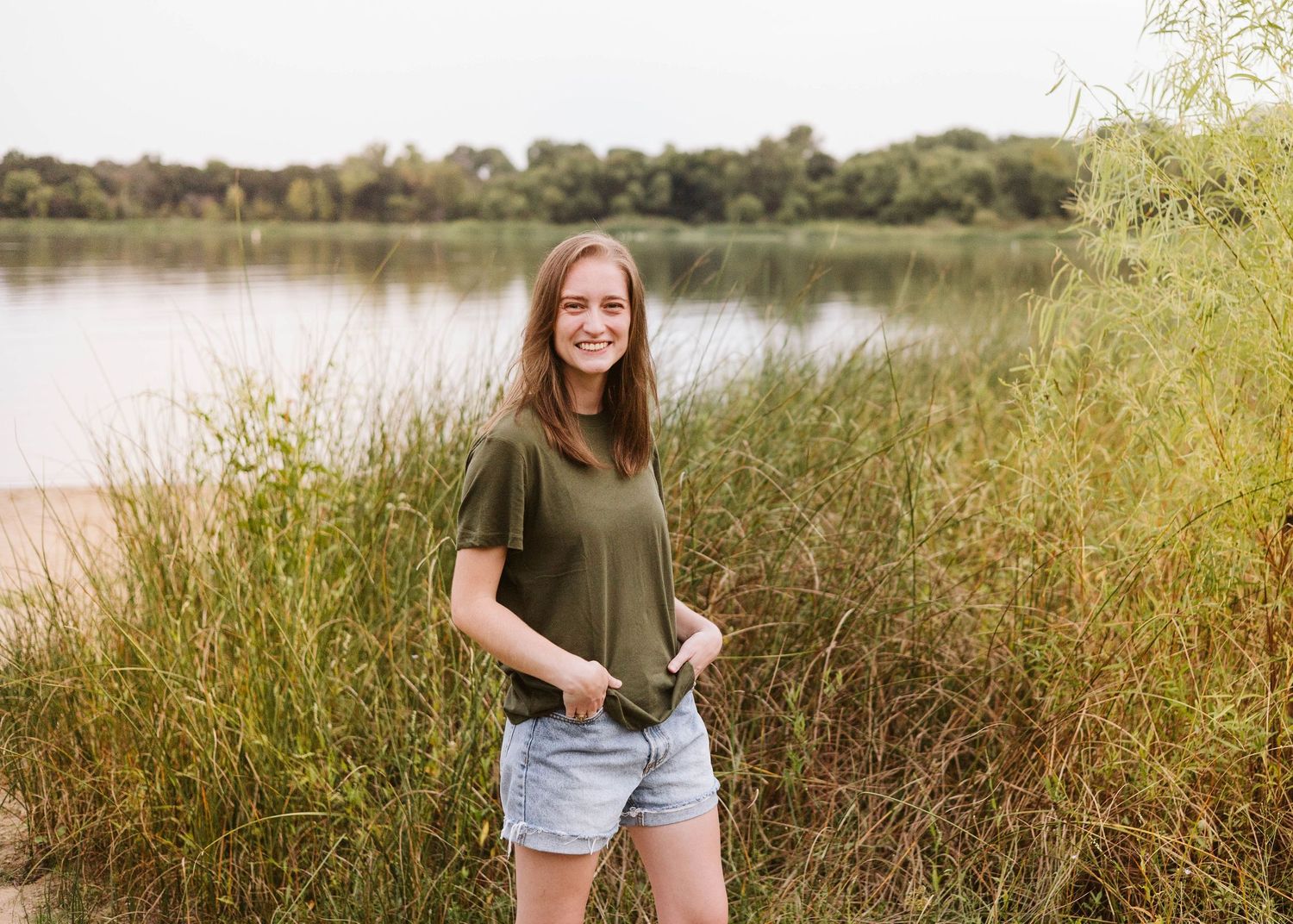 A photo of Brooke, owner of Brooke Gonzalez Photography, standing in tall grass in front of a lake in Fort Worth, Texas on a sunny day.