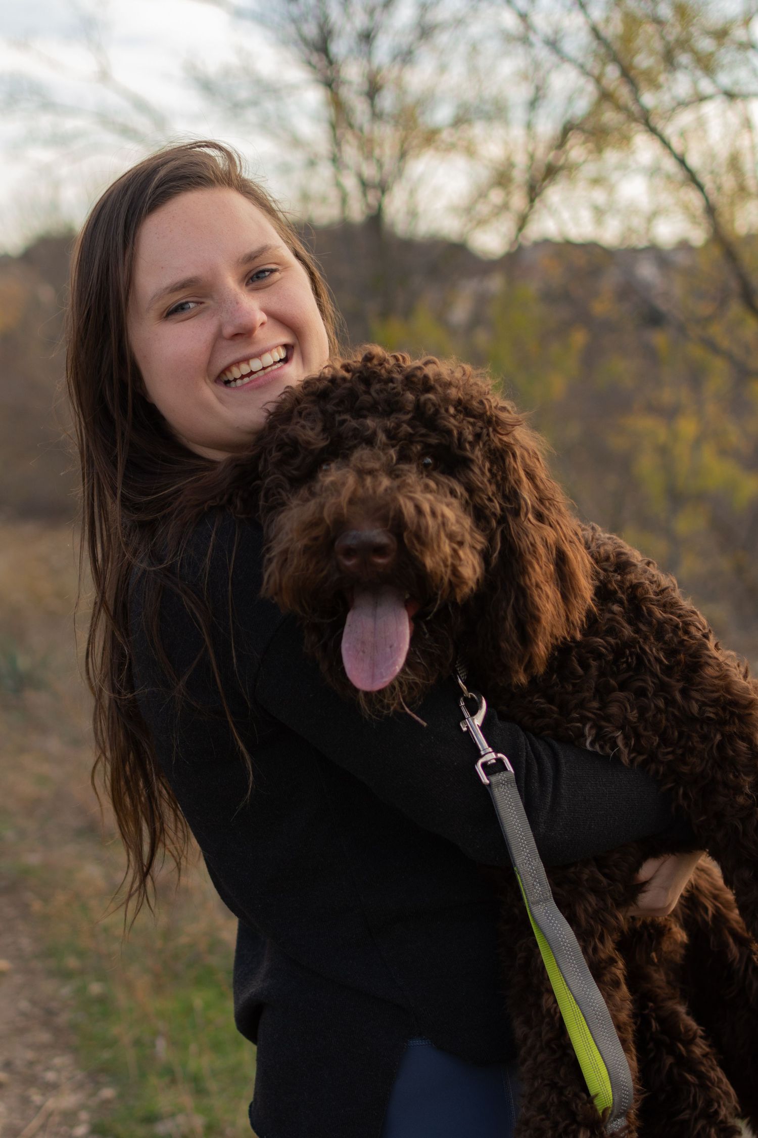 A woman holds up her goldendoodle during a portrait session in Fort Worth during the winter. She smiles as her dog sticks his tongue out.