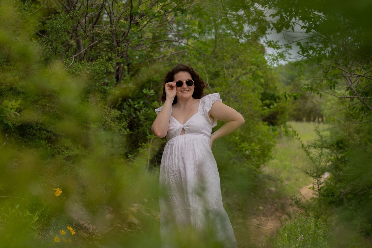 A Masters grad student poses in the trees while wearing a white sundress and touching her sunglasses in Fort Worth during a grad session.