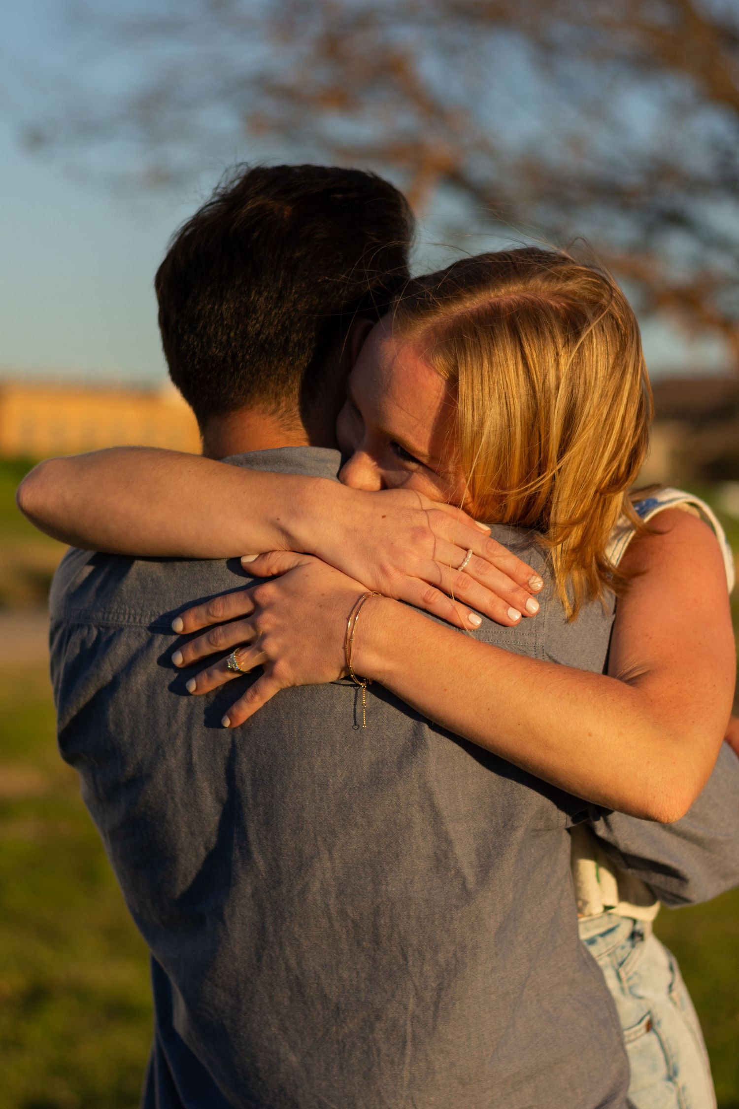 A newly engaged young couple hugs tightly at sunset, full of excitement and emotion, her new engagement ring on her hand on his back.