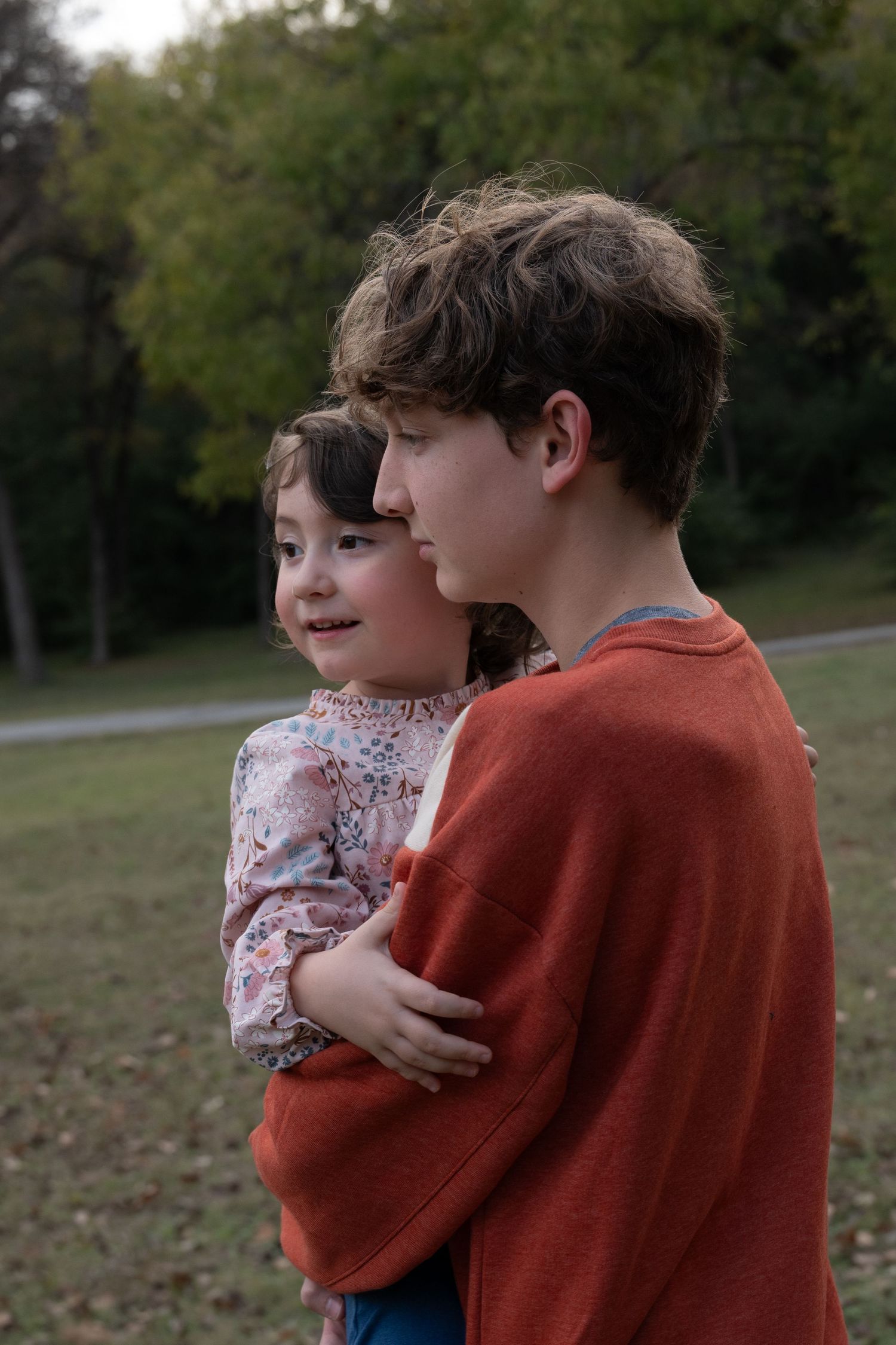Candid of a teen boy holding his little sister as they walk to the next photo spot during a family portrait session in a park in Fort Worth.