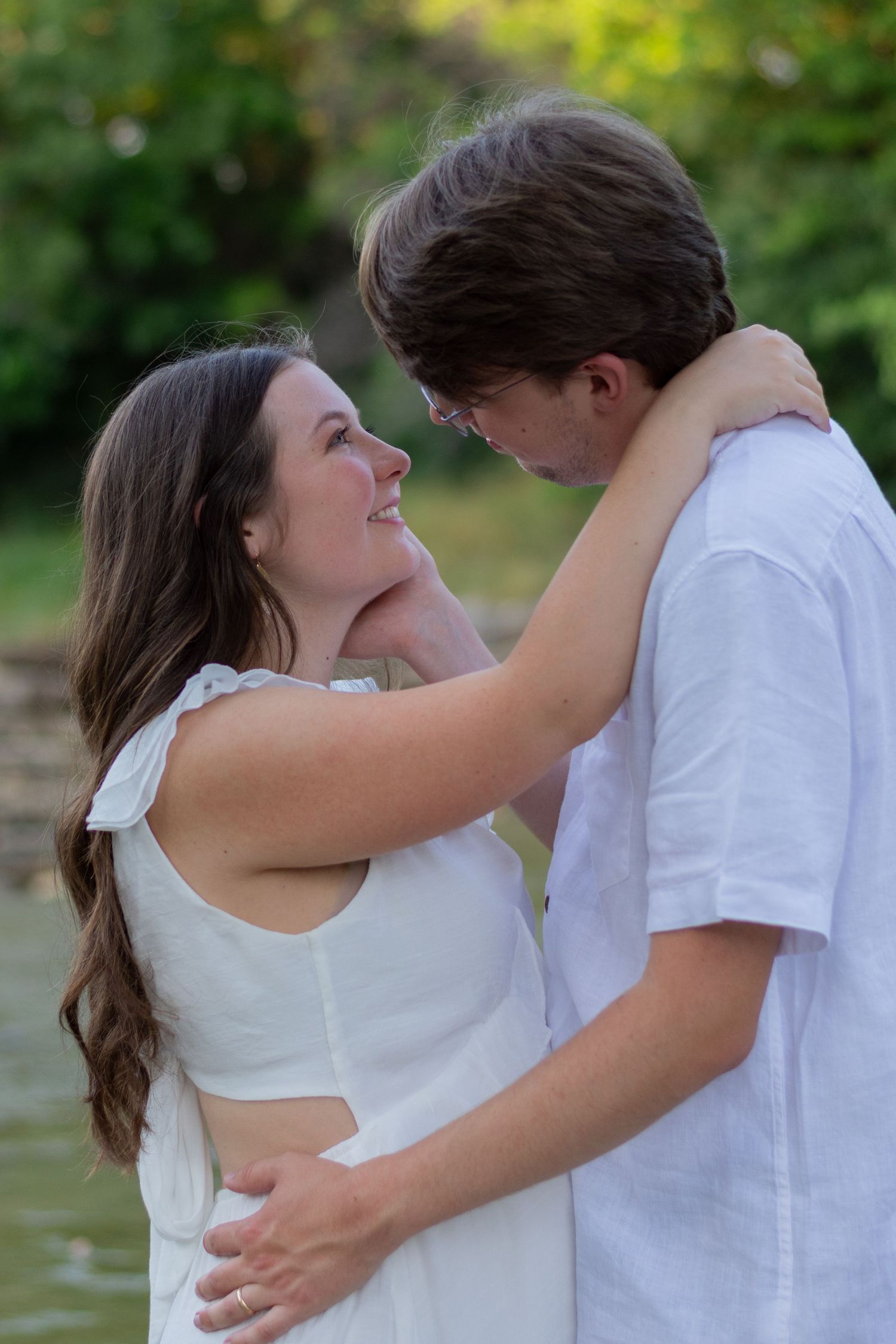 A man looks longingly into his pregnant wife's eyes, holding her face in one hand & waist in other. She smiles back at him with pure love.