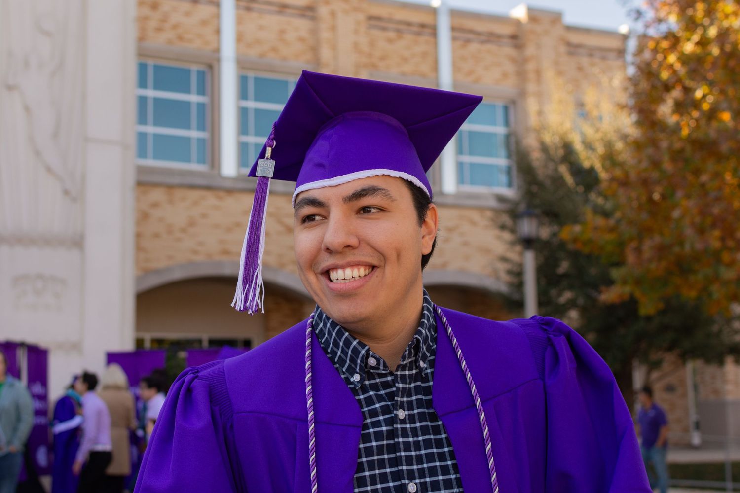 A TCU grad smiles at his family before his graduation ceremony. He has on a plaid shirt, purple cap and gown, and purple and white chord.