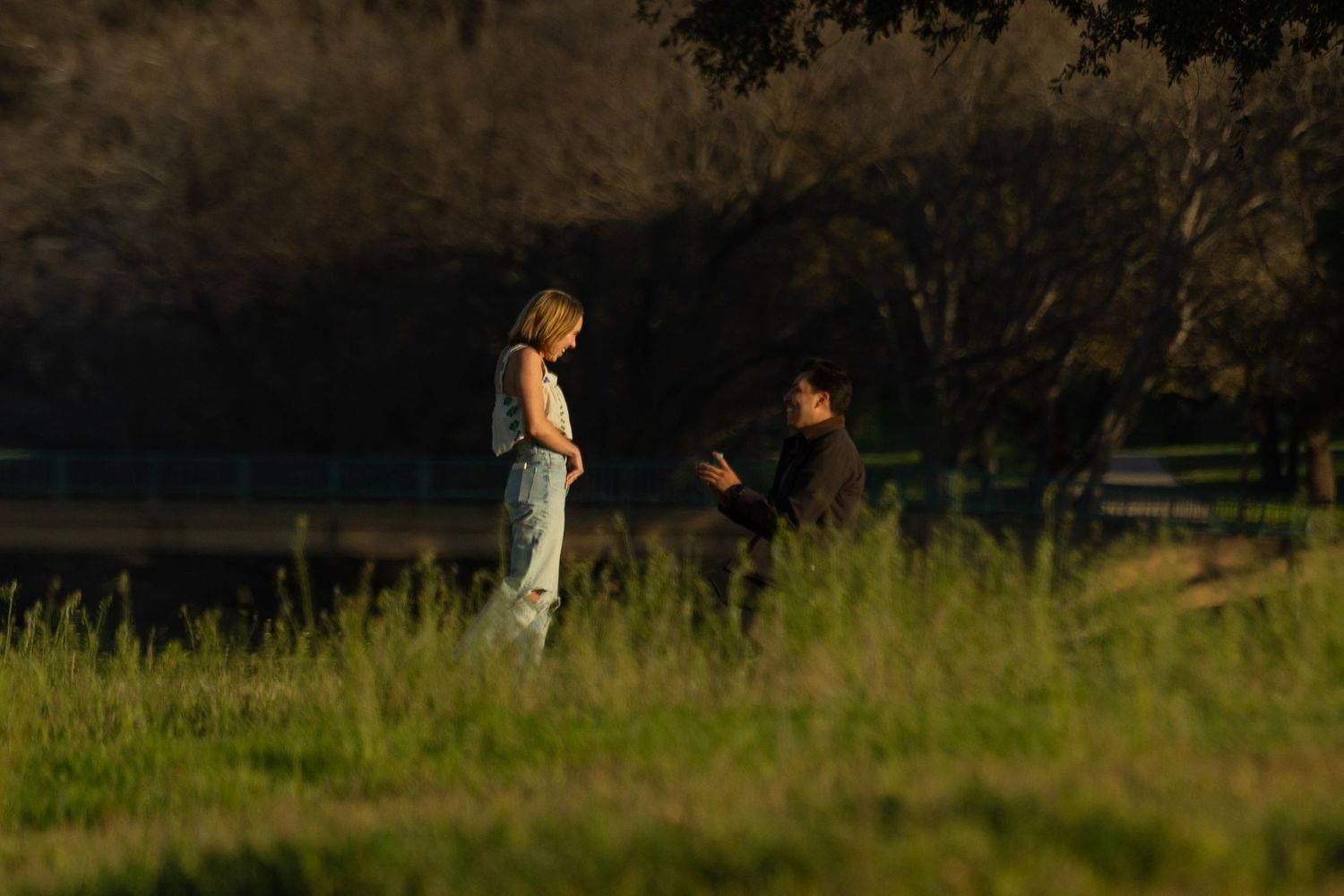 A man kneels on one knee, proposing to his girlfriend at a Fort Worth park at sunset. She inches closer in disbelief and joy to see the ring