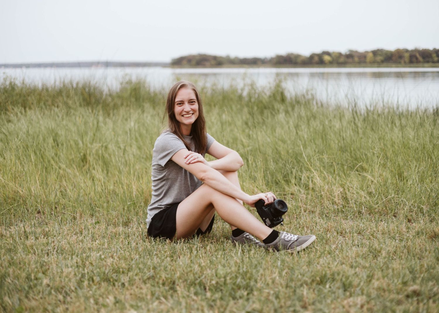 Photo of photographer Brooke sitting in grass in front of a Fort Worth lake as she holds her camera down and smiles on a sunny spring day.