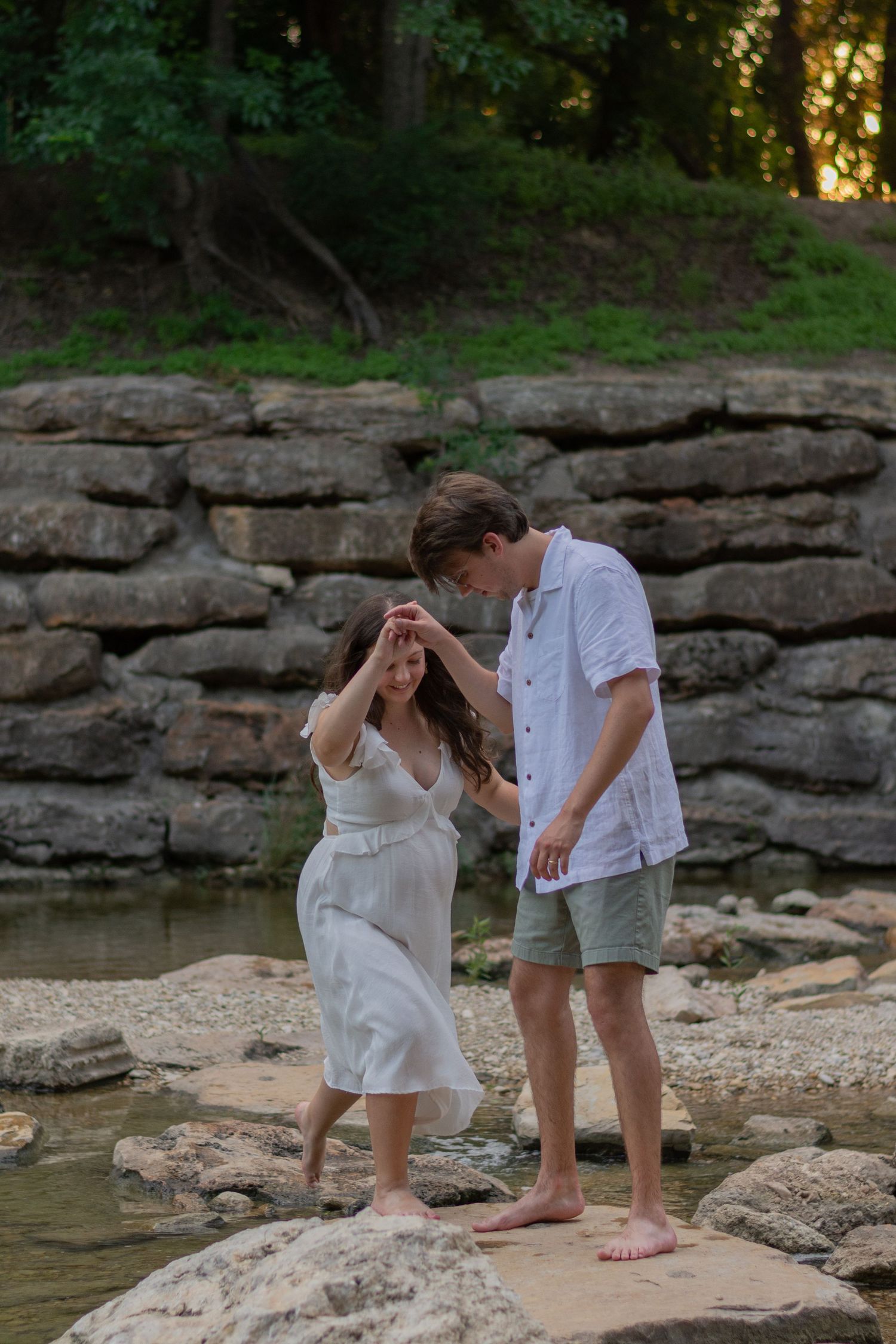 A man helps his pregnant wife across some slippery river rocks during an adventurous summer maternity session in Panther City, Fort Worth.