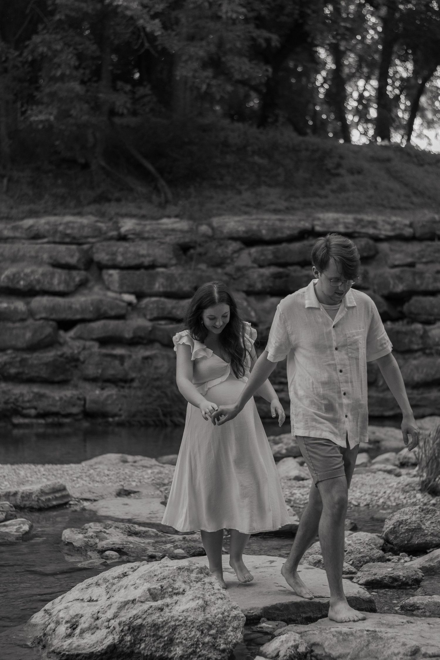 A black and white photo of a couple walking across river rocks while the husband leads his wife, holding hands during a maternity session.