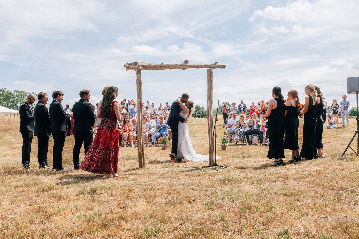 A panoramic view of wedding guests gathered on a grassy hillside under a wooden arch for an outdoor ceremony.