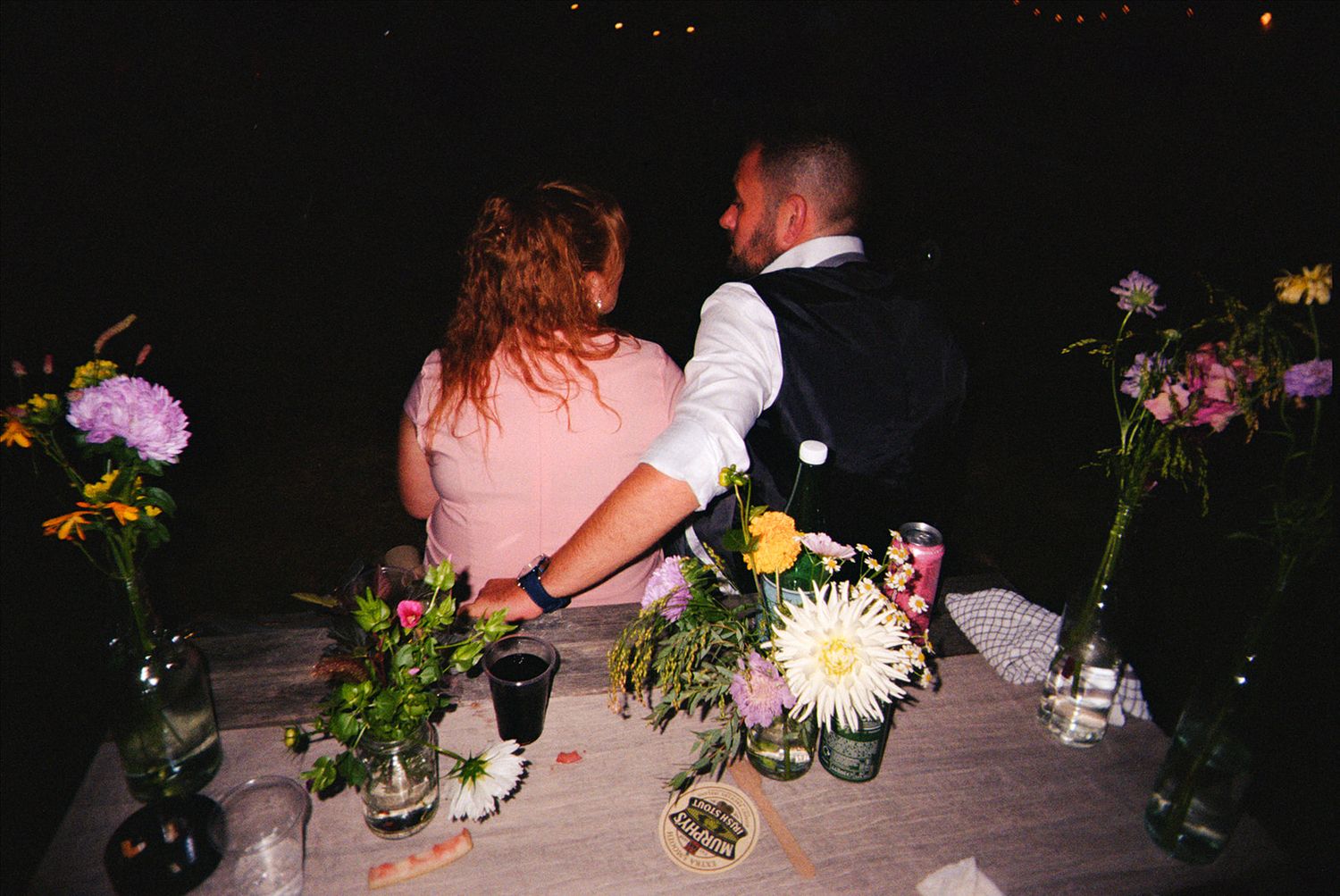 A row of colorful floral centerpieces with candles arranged on a dark wooden table at night.