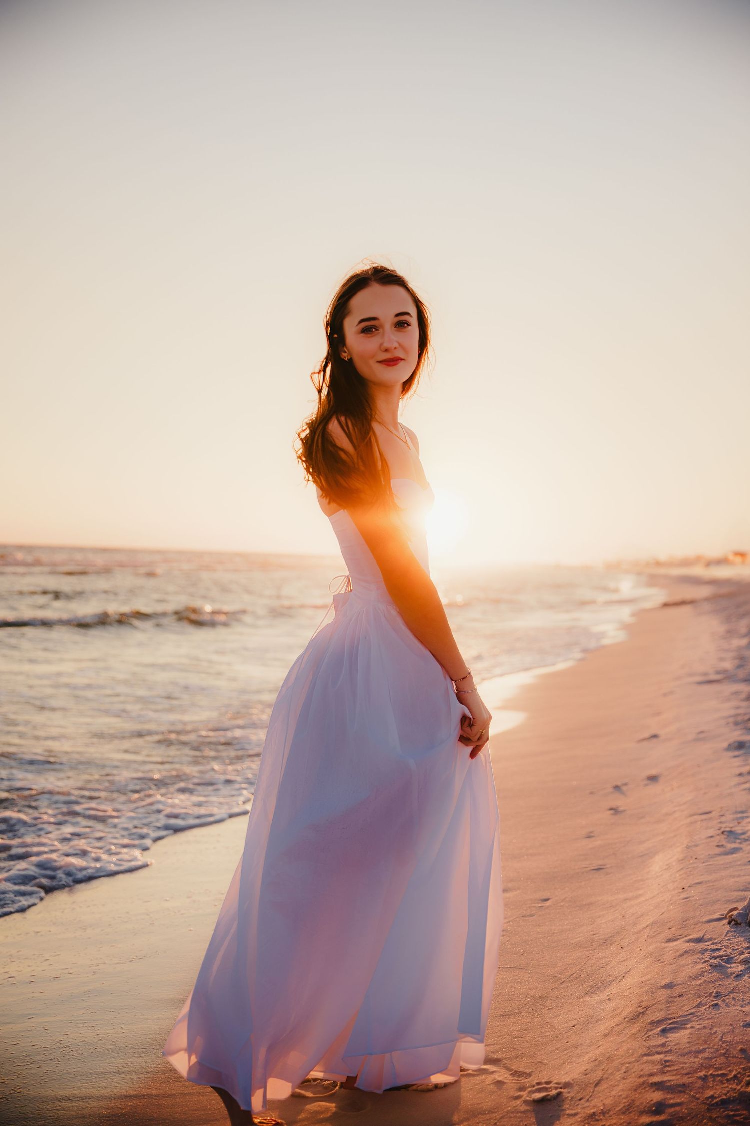 Person in flowing white dress standing on beach during sunset with ocean waves.