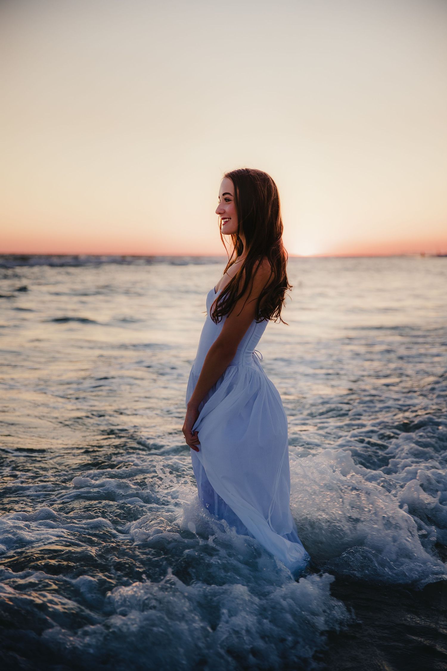 Person wearing white dress standing in ocean waves during sunset.