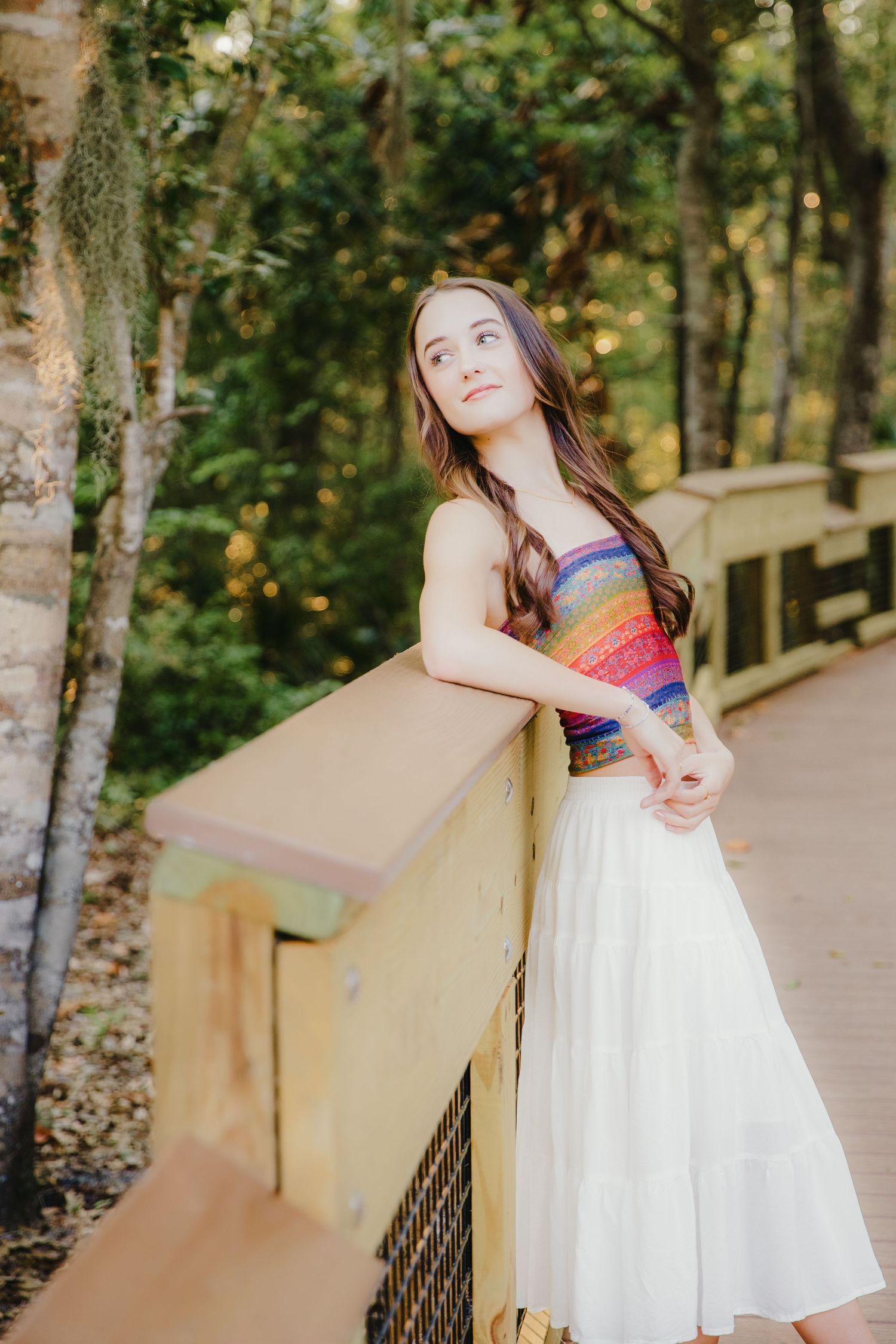Person in rainbow striped top and white skirt leaning on wooden railing in forest setting.